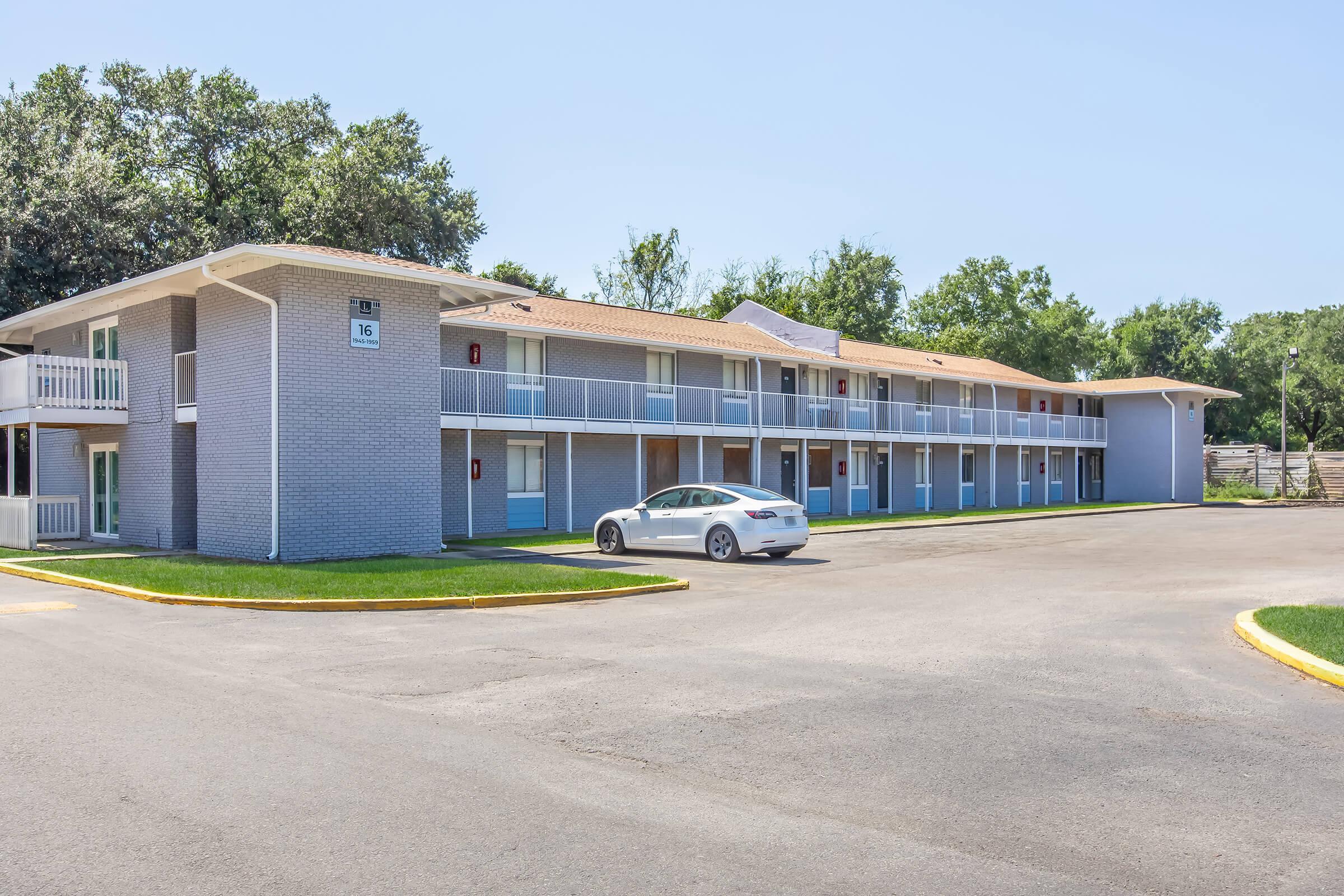Two-story motel building with light gray brick exterior and blue accents. The front features several doors leading to rooms, with a white car parked in the lot. Surrounding the building are green lawns and trees under a clear blue sky.