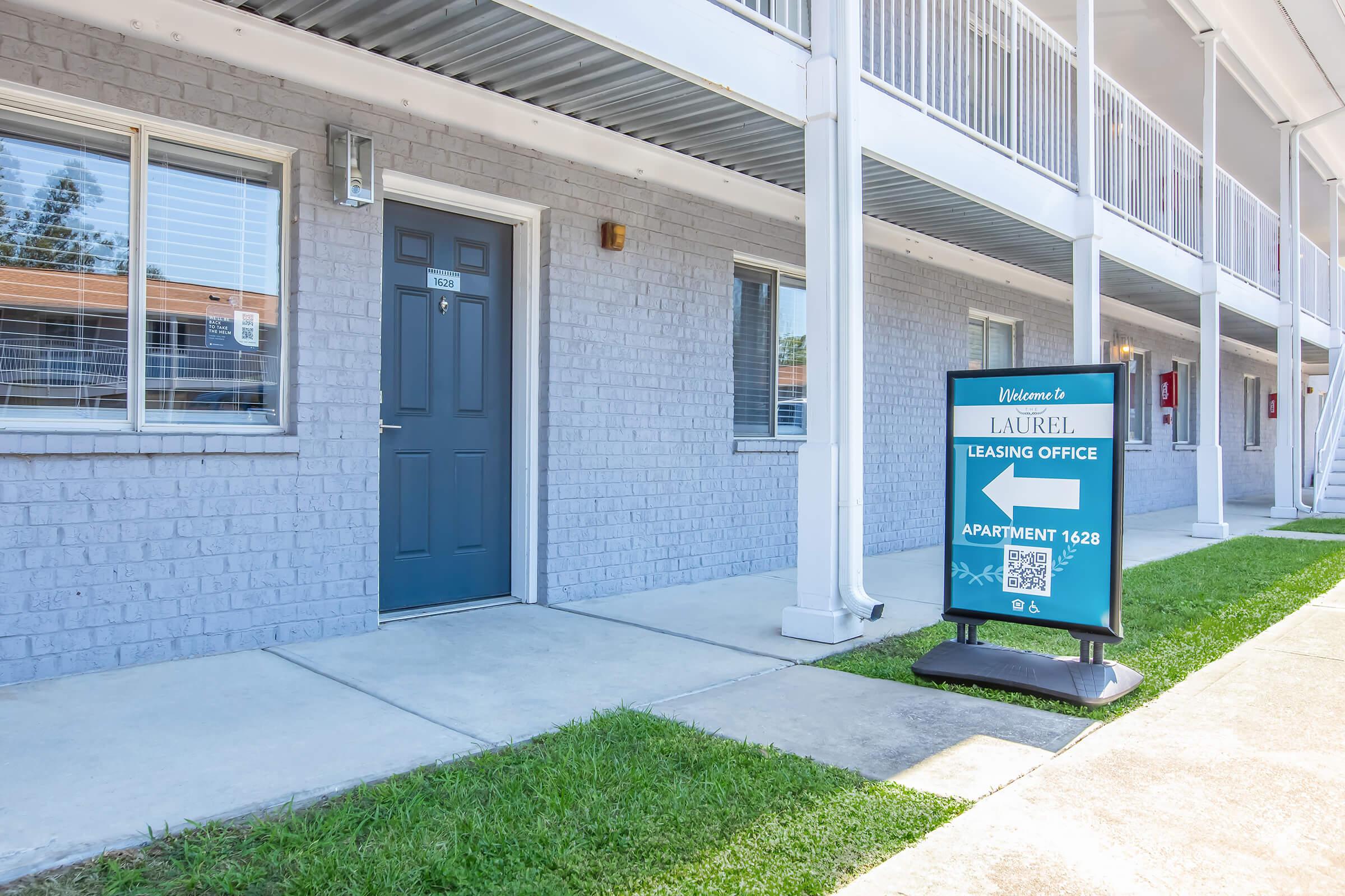 A well-maintained apartment complex exterior featuring a blue door with a welcome sign, a leasing office sign for "Laurel," and an apartment number displayed. The building has several windows, is painted in light gray, and has a grassy area in front.