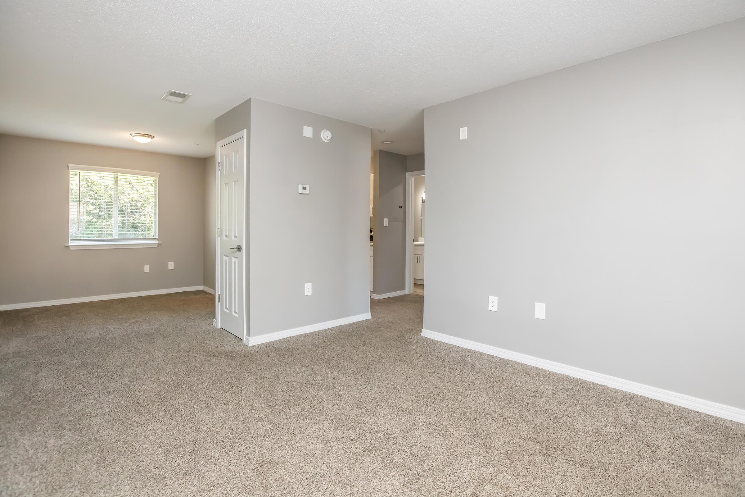 A spacious, empty room with light gray walls and carpeted flooring. Sunlight streams through a window on the left, illuminating the space. A doorway leads to a bathroom in the background, and a closet is visible on the right. The overall atmosphere is clean and airy, suitable for various uses.