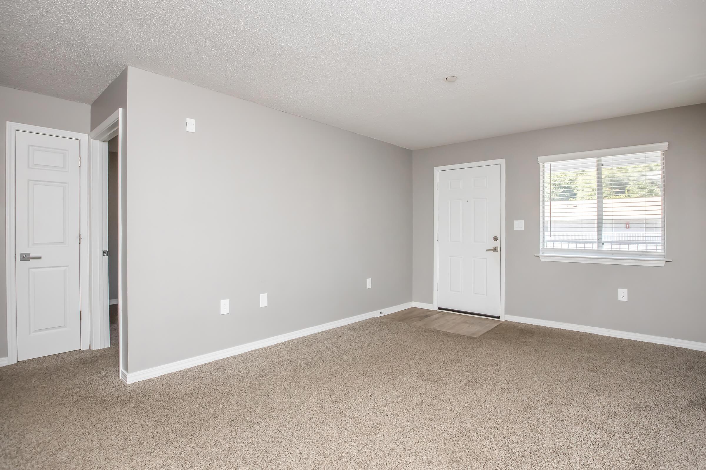 Empty room with light gray walls and a tan carpet. There is a white door leading outside, a window with blinds allowing natural light, and a closet door visible on the left side. The space is well-lit and feels open and inviting.