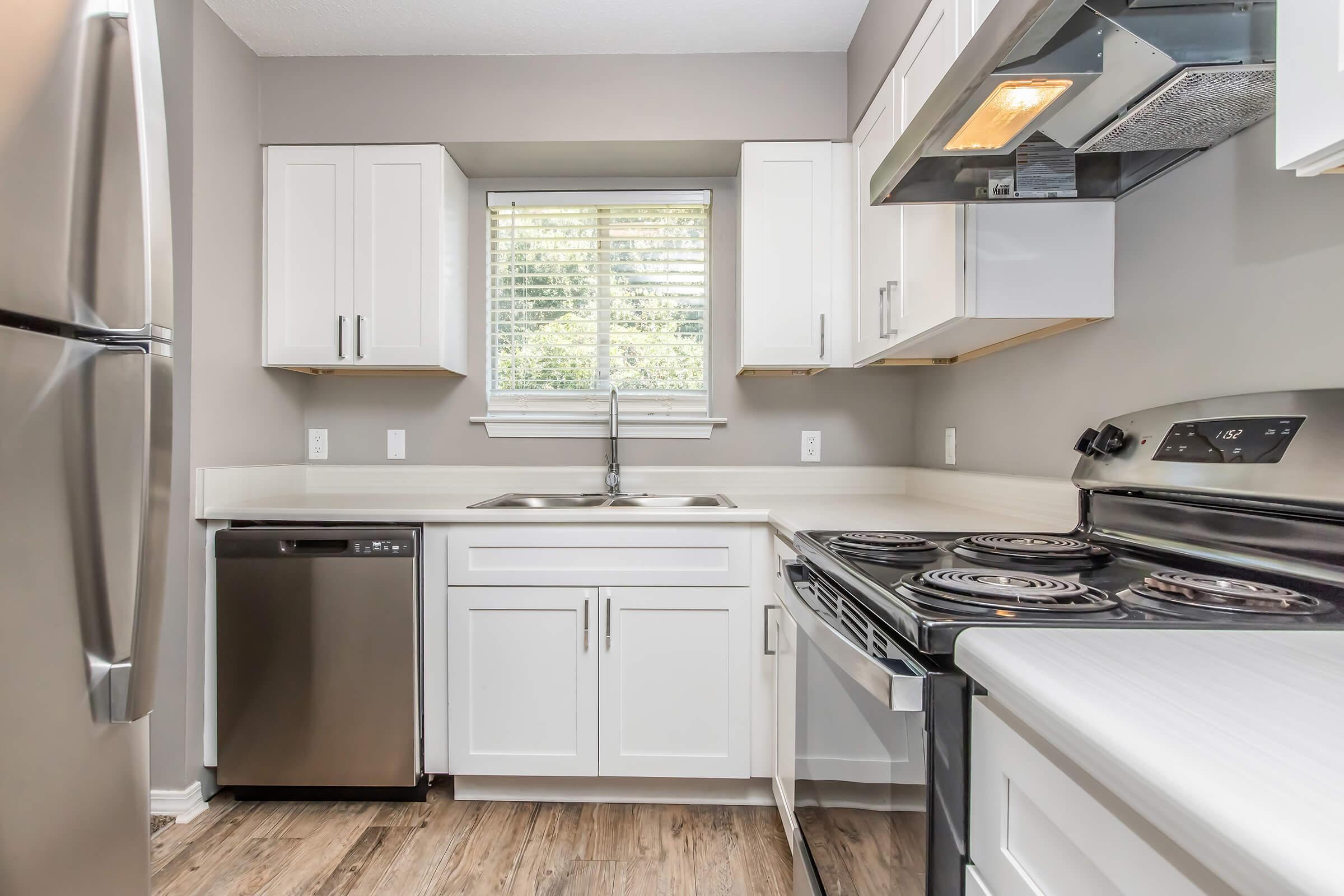Modern kitchen featuring stainless steel appliances including a refrigerator and dishwasher. White cabinetry with silver handles, a double sink beneath a window, and a stove with an oven. Light-colored countertops and wooden flooring complete the contemporary design.