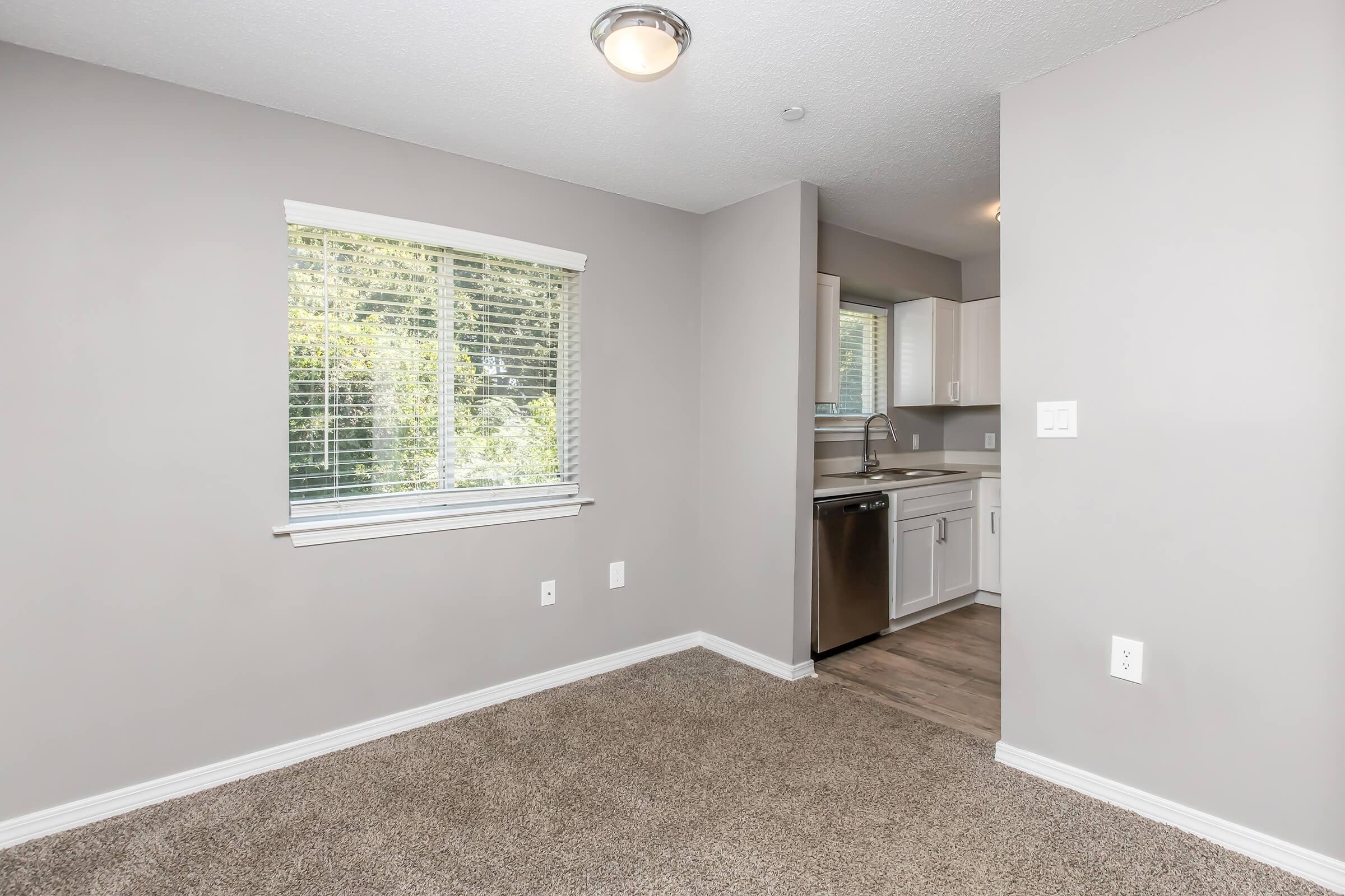 A well-lit interior of a small room featuring beige carpet, a window with vertical blinds, and a view of greenery outside. The adjacent kitchen area has white cabinets and stainless steel appliances, including a dishwasher. The walls are painted a light gray, creating a modern and inviting atmosphere.