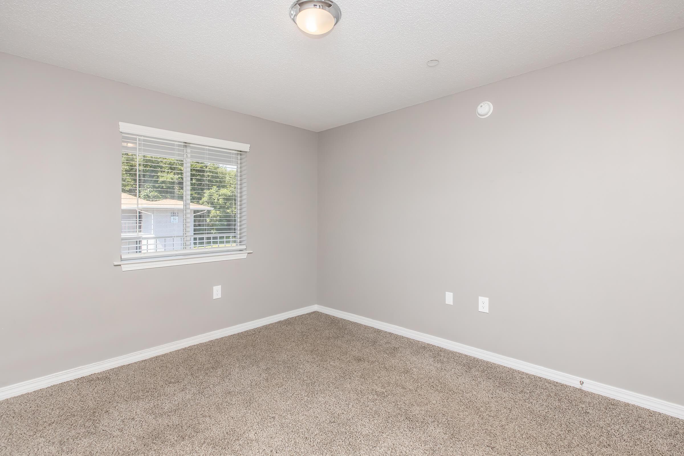 Empty room with light gray walls and carpeting. A small window with white blinds allows natural light to enter, illuminating the space. There is a ceiling light fixture, and the room features two electrical outlets on the wall. No furniture or decorations are present, creating a minimalist appearance.