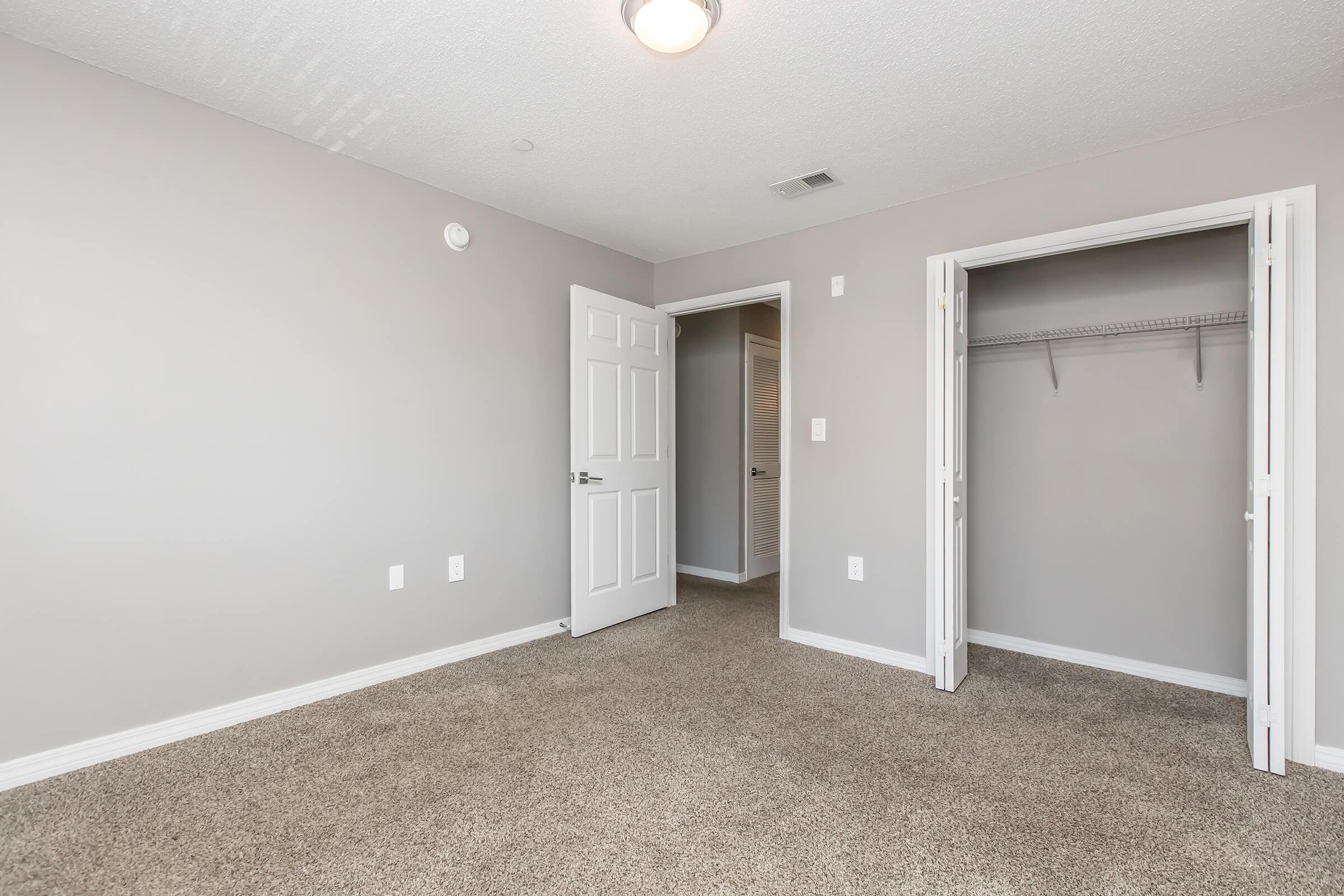 A spacious, empty room with light gray walls and beige carpeting. There is a closed door on the left leading to another room, a closet with a shelf open on the right, and a ceiling light fixture providing illumination. The overall space is well-lit and minimalistic, suitable for various purposes.