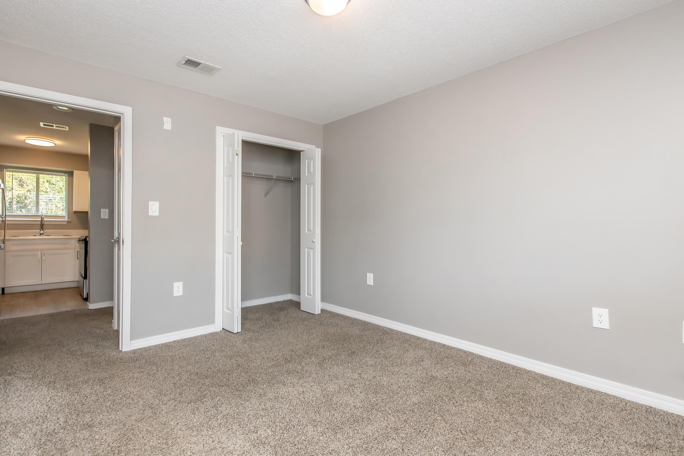 Empty room with light gray walls and carpeted floor. Two doors lead to closets and a bathroom area in the background. A ceiling light fixture is visible, illuminating the space. The room is well-lit with natural light coming from the doorways.