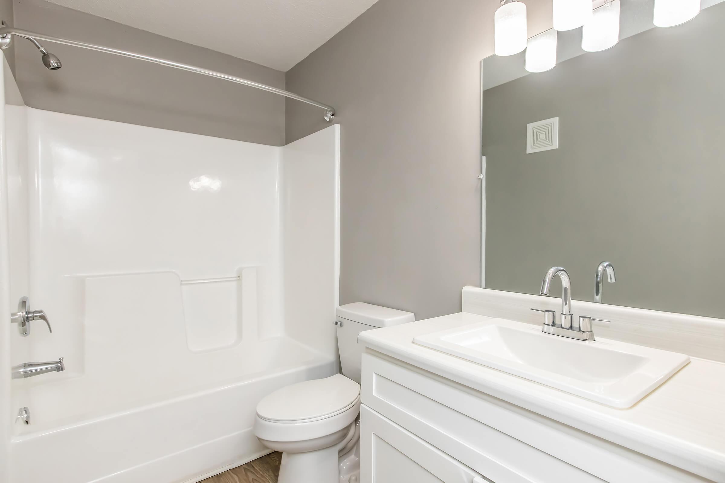 A modern bathroom featuring a white bathtub with a shower rod, a clean white toilet, and a sleek vanity with a white sink and faucet. The walls are painted in a muted gray color and illuminated by overhead light fixtures.