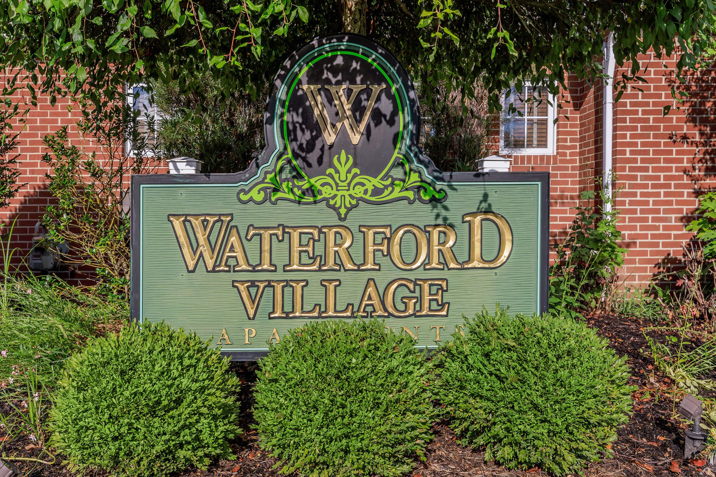 Sign for Waterford Village Apartments displaying the name prominently in gold letters against a green background, surrounded by decorative elements and lush greenery.