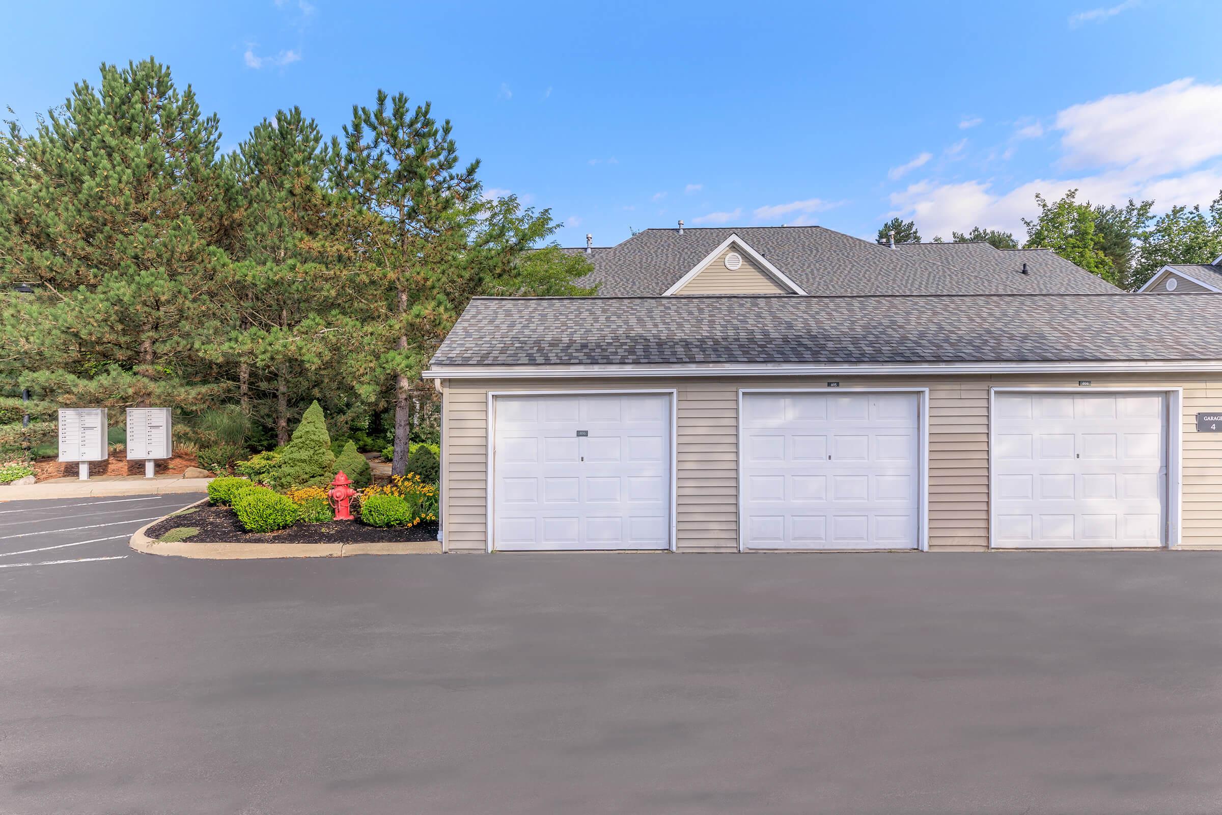 Two garage doors in a residential area, flanked by green bushes and trees. In the background, there are additional storage units or garages. The asphalt driveway is visible, and the sky is clear with a few clouds. The area is well-maintained and features landscaping.