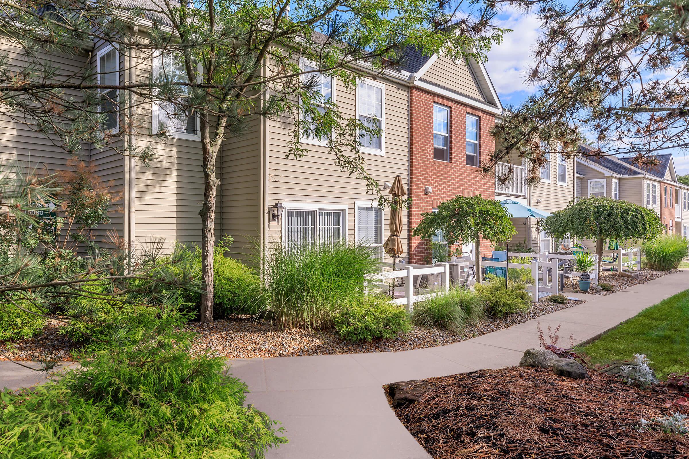 A well-maintained residential area featuring two-story apartment buildings with a mix of brick and siding exteriors. Lush greenery, including shrubs and trees, line the walkway, which leads to outdoor seating areas with patio furniture. The scene is bright and inviting, under a clear blue sky.