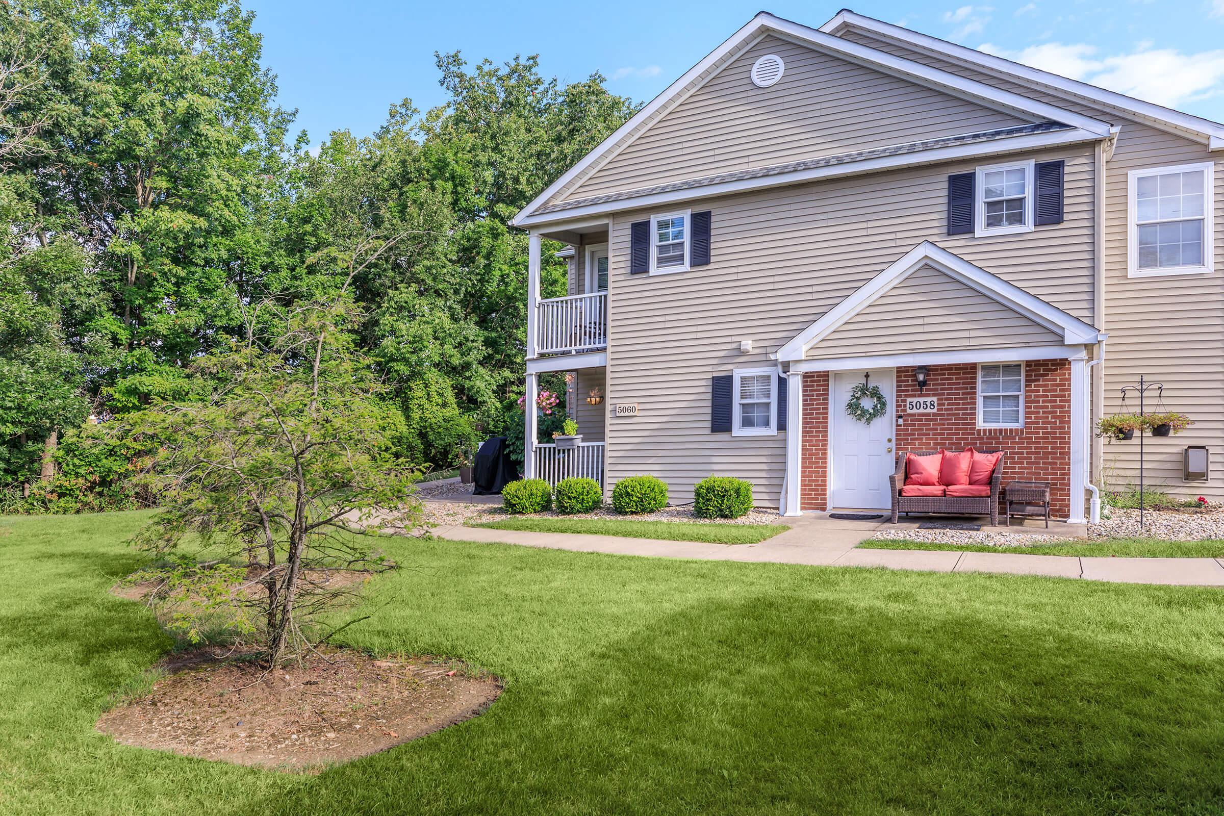 A two-story beige house with a porch and a welcoming red bench. Lush green grass surrounds the property, along with neatly trimmed bushes in front. Trees and greenery line the background, providing a serene and inviting atmosphere.