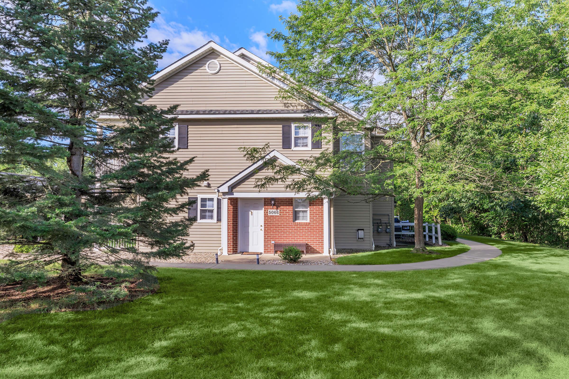 A two-story residential house with a light-colored exterior, red brick accents, and a well-kept lawn. Surrounding the house are trees and shrubs, with a paved path leading to the entrance. The sky is blue with a few clouds, creating a bright and inviting atmosphere.