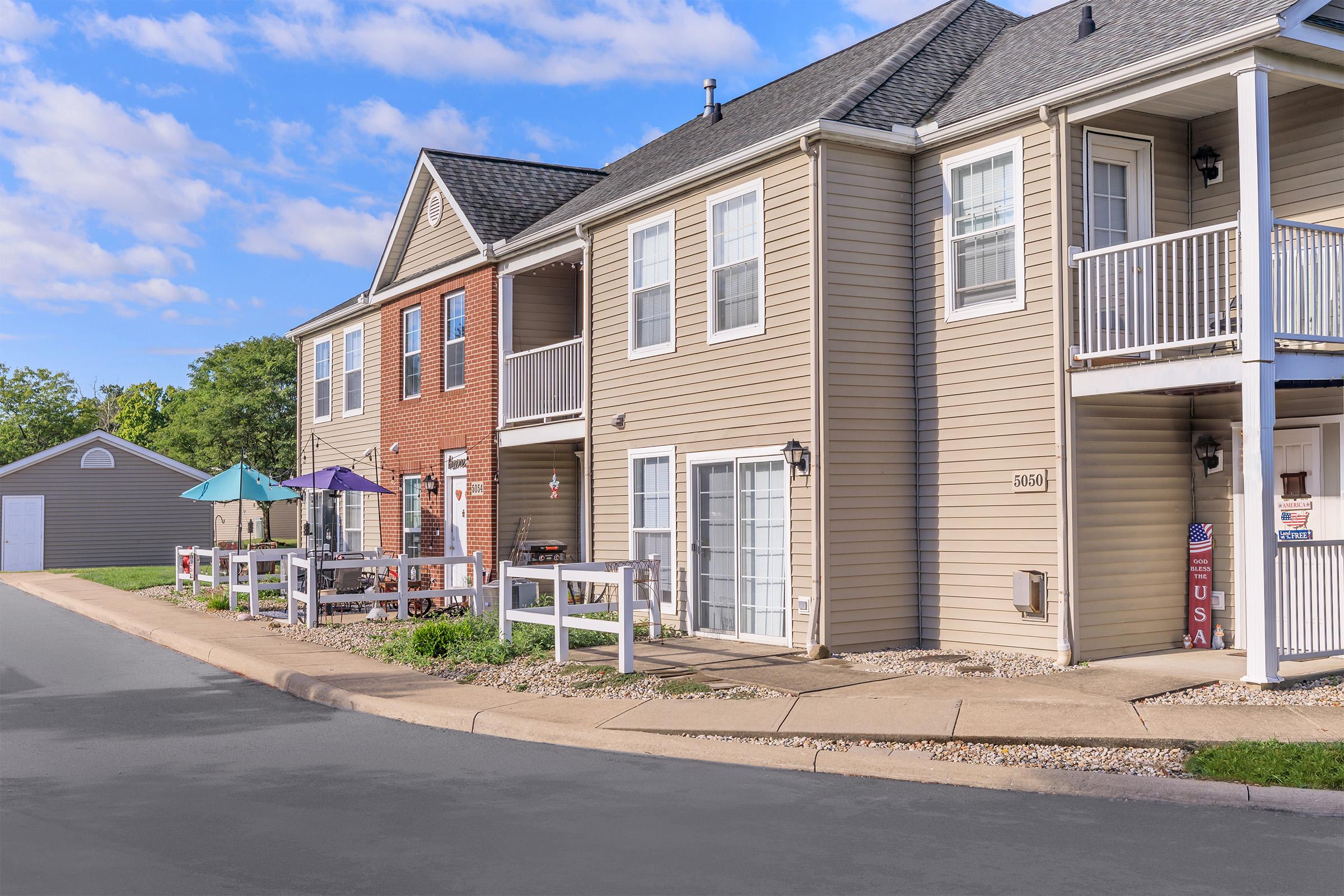 A residential building with a mix of beige and brick facades. The front features a concrete walkway, small porches with umbrellas, and white railing. There are several windows and a neatly maintained landscape. In the background, there's a garage and blue sky with scattered clouds.