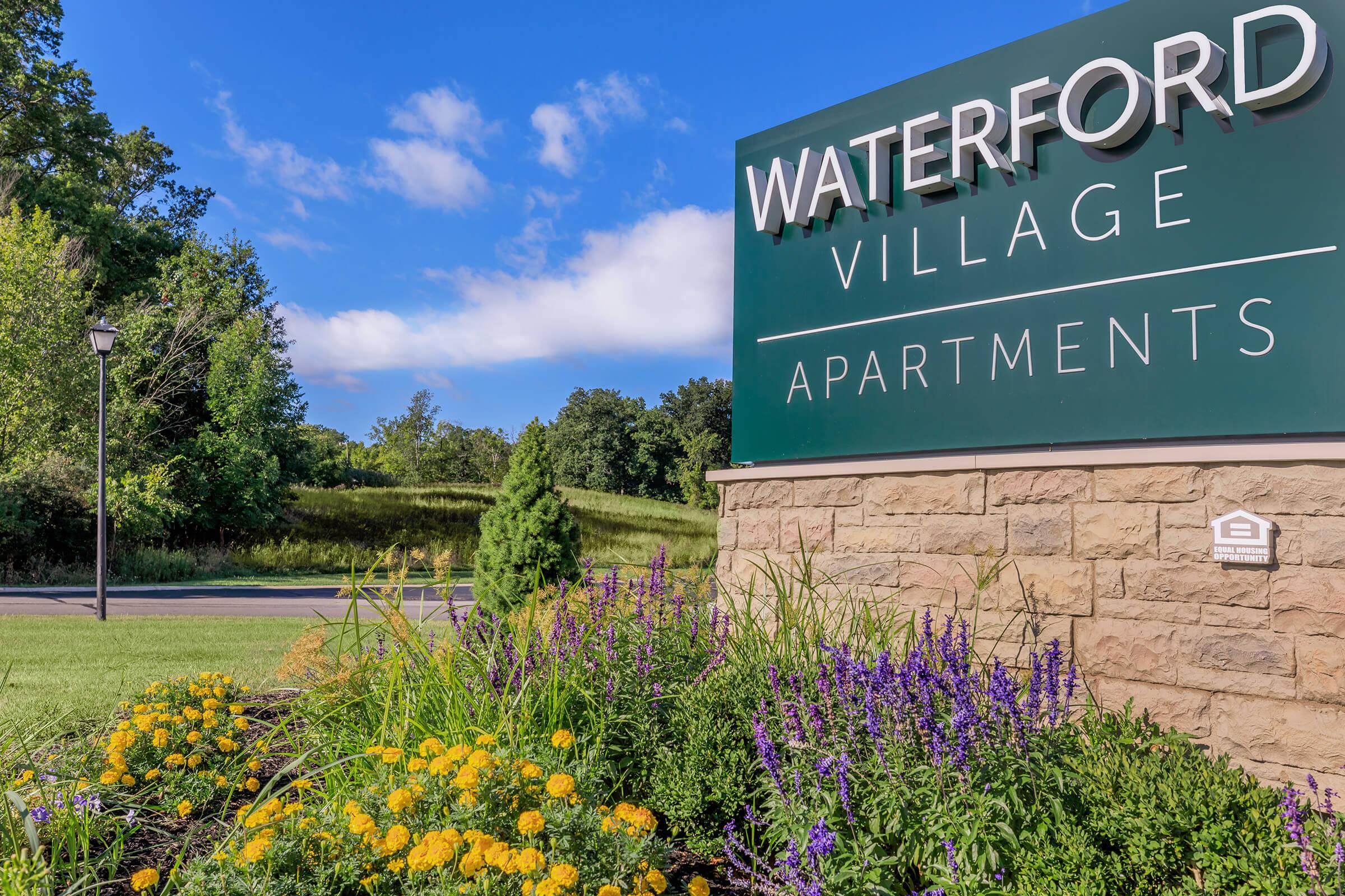 Sign for Waterford Village Apartments with a landscaped garden featuring colorful flowers, set against a clear blue sky and green trees in the background.