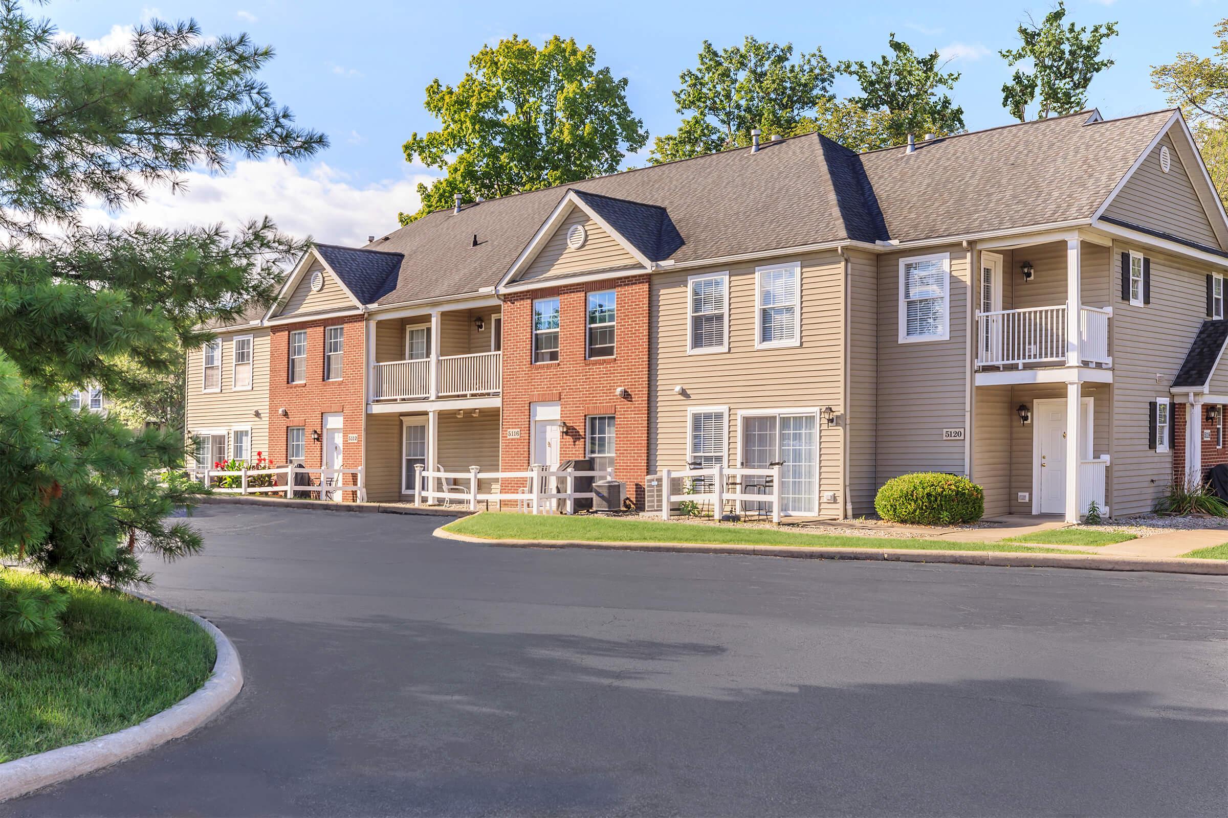 A residential property featuring two-story apartment buildings with brick and beige siding, surrounded by green grass and trees. The driveway is paved, leading to the entrance of the buildings. Clear blue sky and sunlight illuminate the scene, giving a welcoming and well-maintained impression.