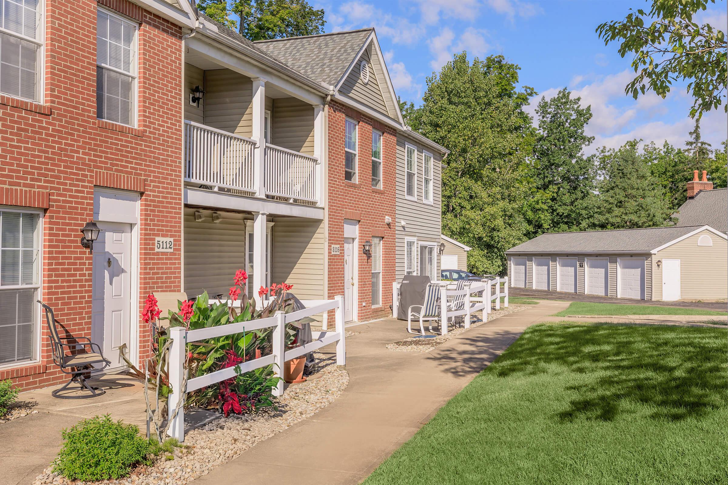 A landscaped walkway leads to a two-story brick apartment building with white balconies. Flower beds with colorful plants are in the foreground, and a series of storage garages are visible in the background under a clear blue sky. Lush green grass surrounds the area, creating a welcoming environment.
