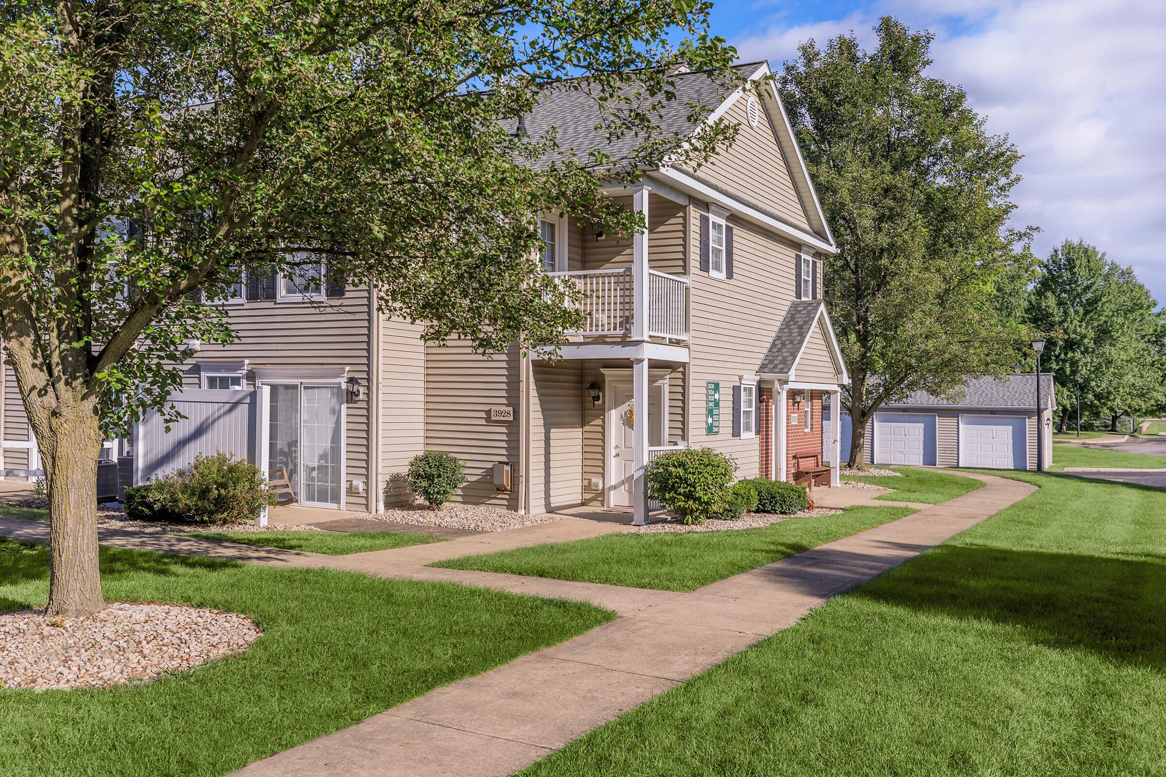 A well-maintained residential building with light brown siding, featuring multiple apartment units. The landscaped area includes manicured lawns, trees, and small bushes. A paved walkway leads to the entrance, while a garage is visible in the background along with a clear blue sky.