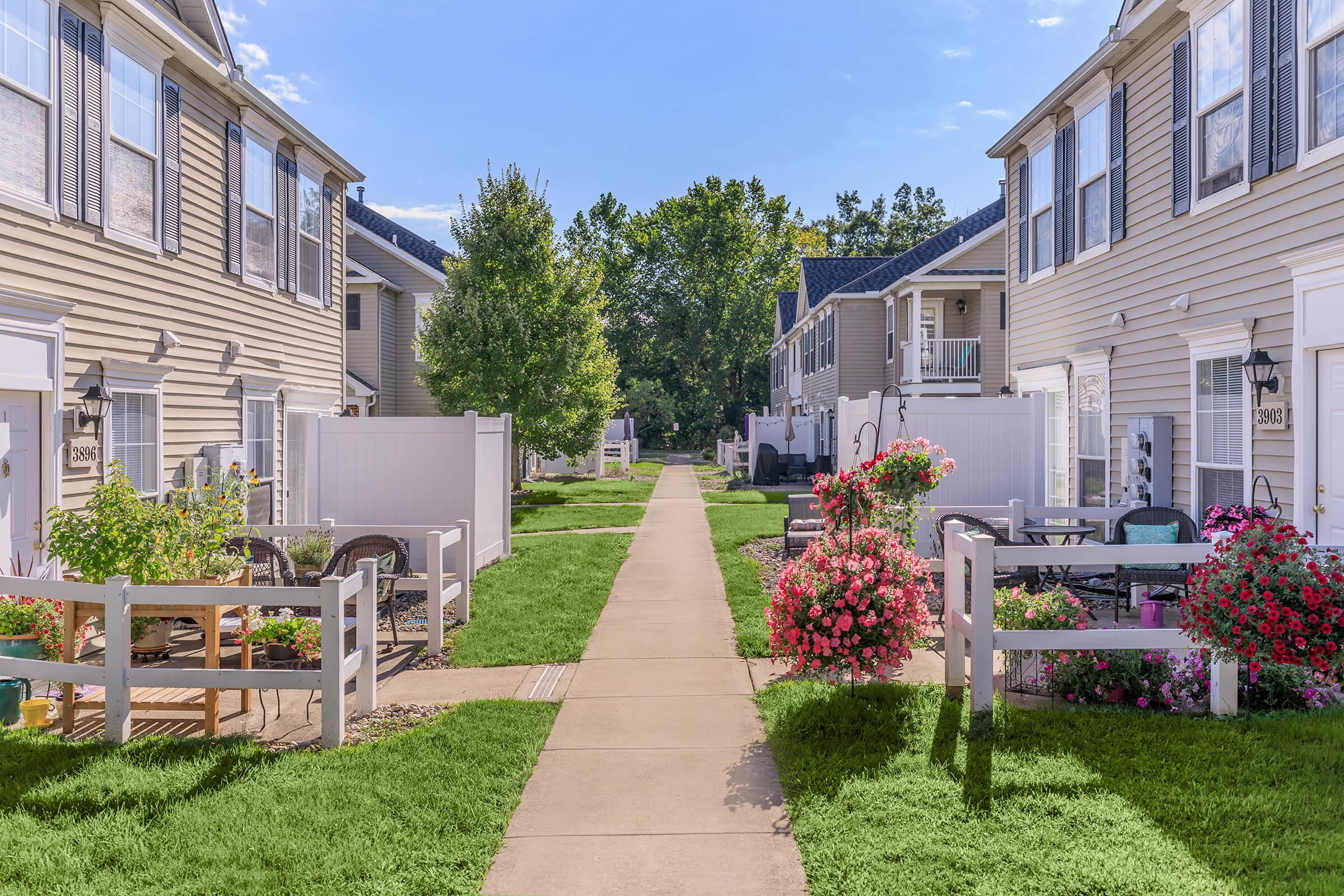 A tranquil residential pathway lined with well-maintained grass and colorful flower beds. Townhouse units with white fences are visible on either side. The pathway leads to a green area with trees, creating a serene neighborhood atmosphere under a clear blue sky.
