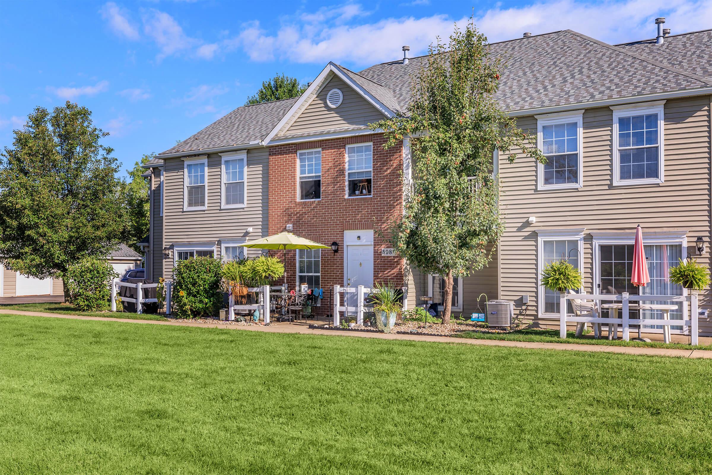 A well-maintained two-story apartment building with light brown siding and a brick accent. A lush green lawn in the foreground, with patio furniture and an umbrella on a small deck. Flower pots add color, and trees line the area. Bright blue sky with a few clouds enhances the inviting atmosphere.