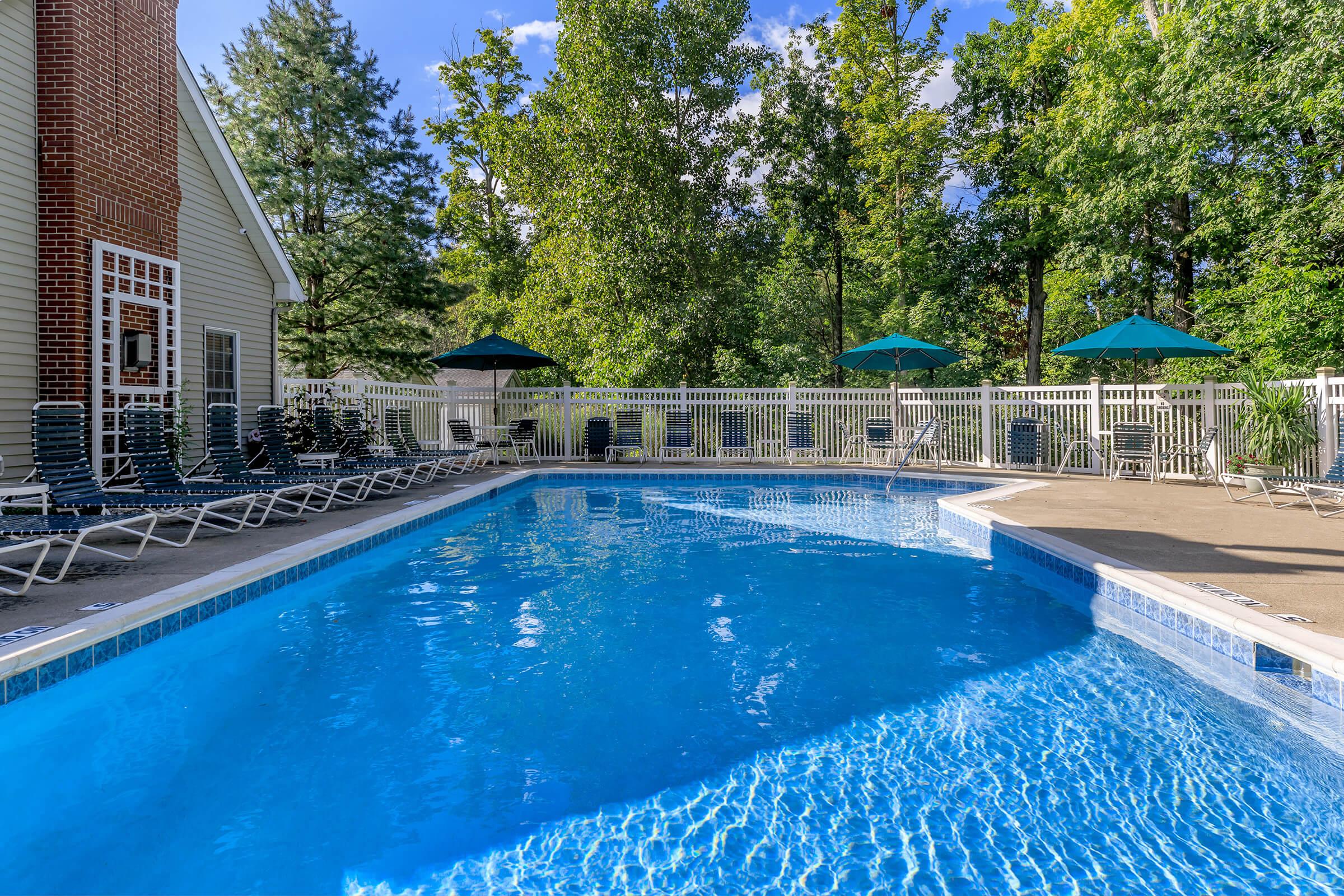 A serene outdoor pool area with clear blue water, surrounded by green trees. Sun loungers are neatly arranged along the poolside, and several umbrellas offer shade. A white fence encloses the area, enhancing the relaxing atmosphere.