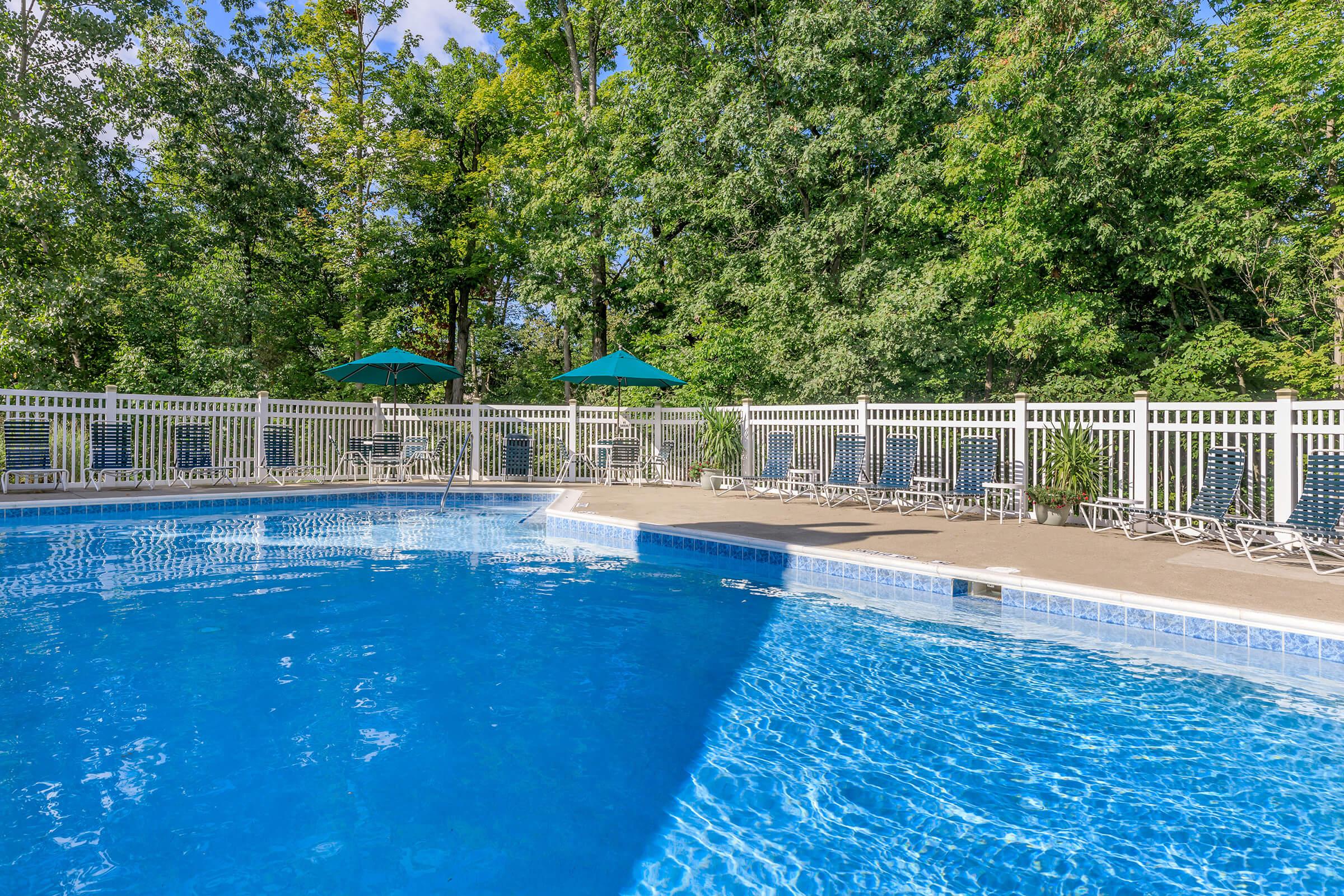 A swimming pool surrounded by greenery, featuring clear blue water, lounge chairs, and two green umbrellas. The area is enclosed by a white picket fence, with trees in the background providing a natural ambiance.
