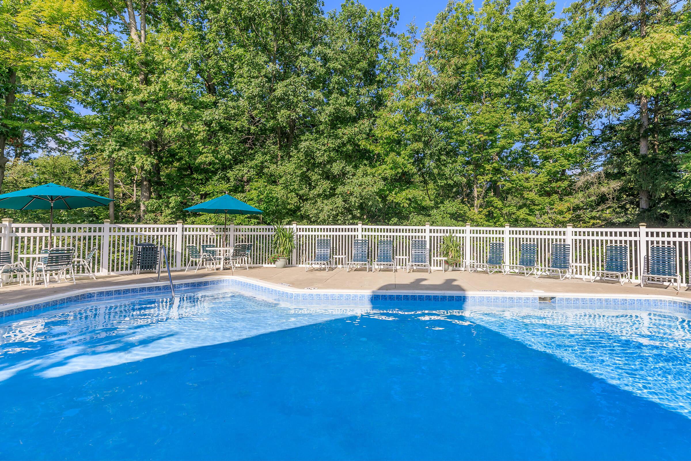 A clear blue swimming pool surrounded by a white fence, with green trees in the background. Several lounge chairs are positioned around the pool, and green umbrellas provide shade. The scene is bright and inviting, reflecting a peaceful outdoor setting ideal for relaxation.