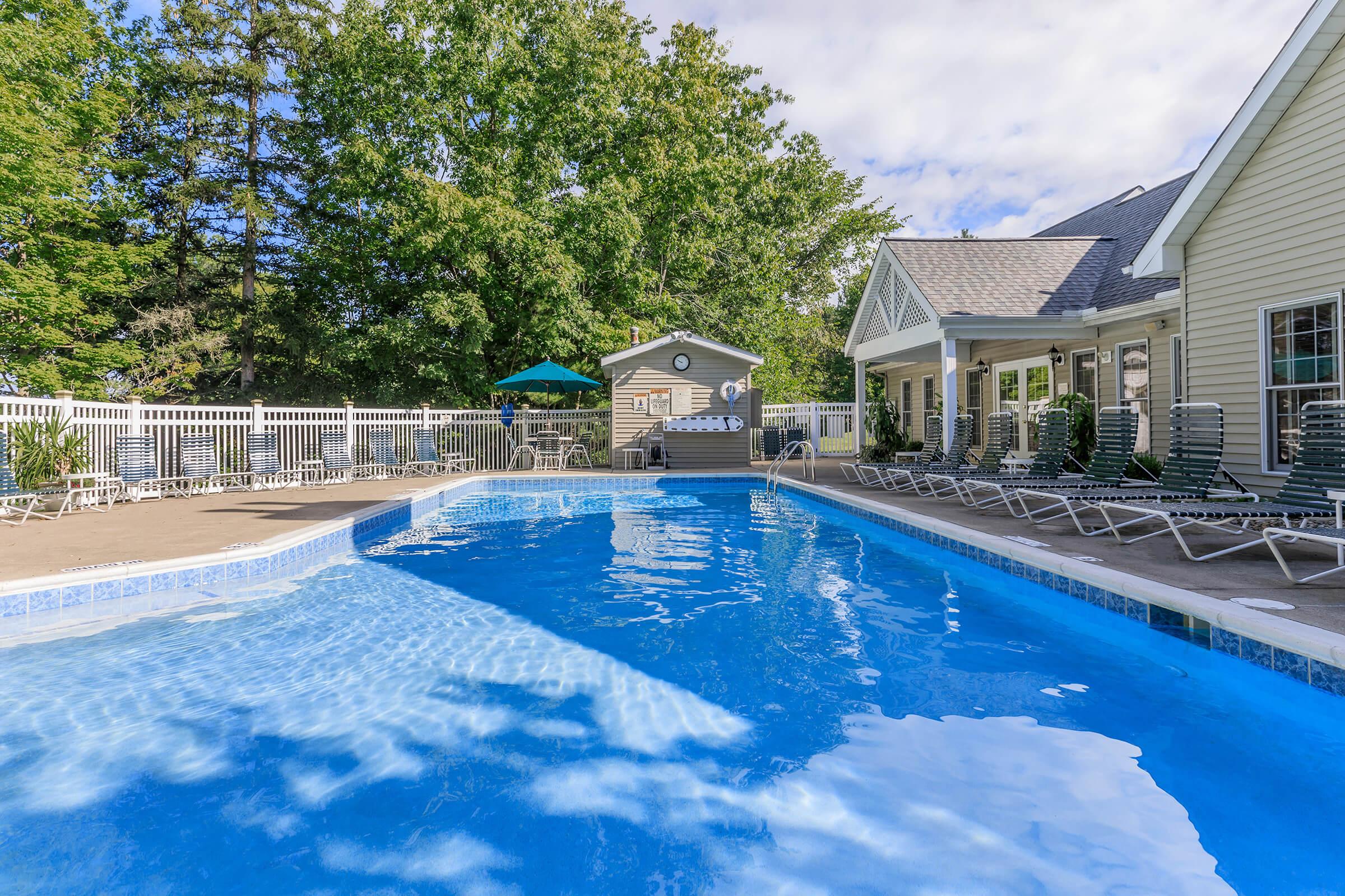 A sparkling blue swimming pool surrounded by lounge chairs and shaded by trees. A small building with a clock is visible in the background, alongside a few umbrellas providing shade. The scene is bright and inviting, perfect for relaxation and leisure.