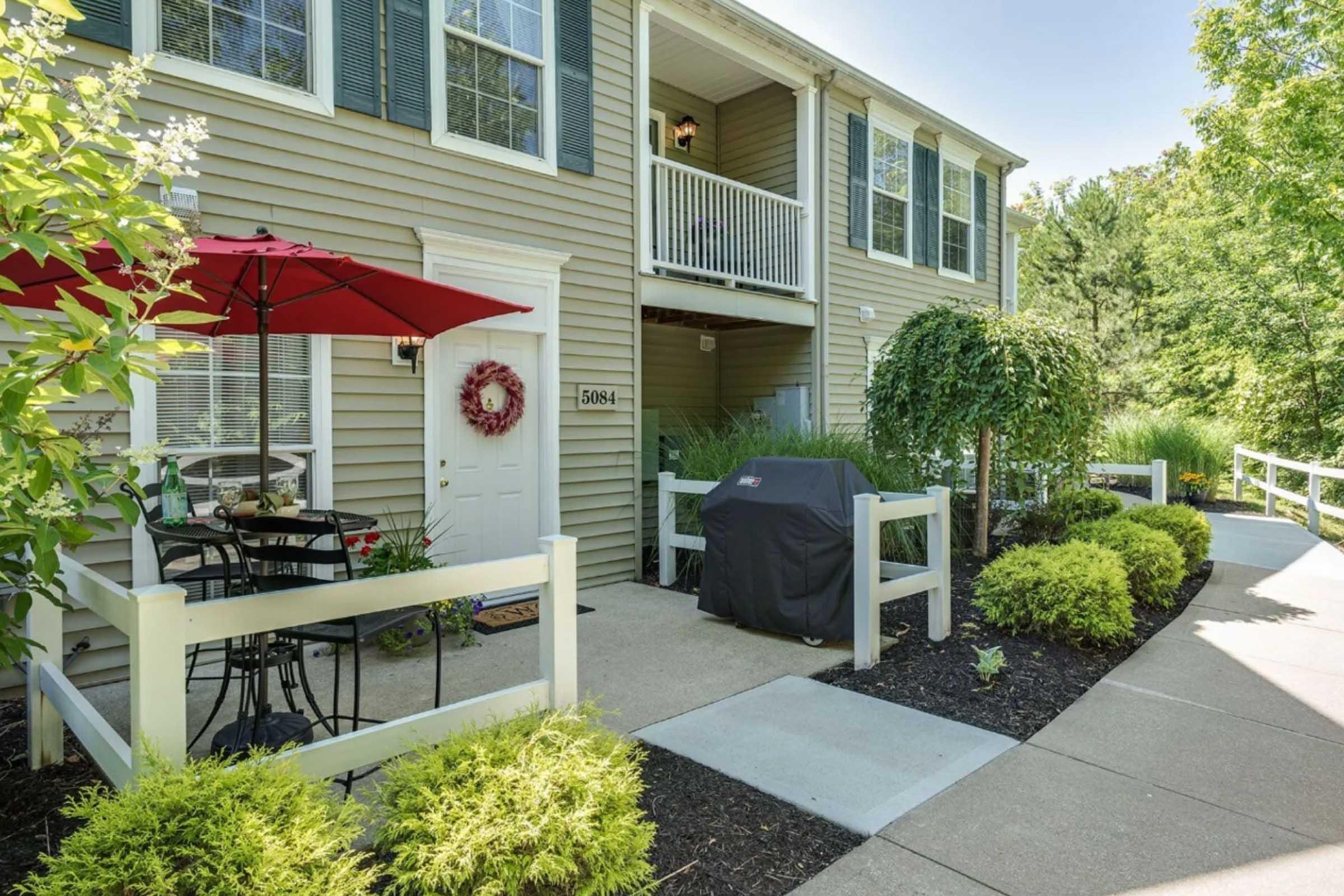 A charming exterior view of an apartment with green siding, featuring a small patio area with a black grill, a round table with chairs, and an umbrella. Lush greenery and flowers line the walkway, leading up to the entrance with a decorative wreath on the door. Sunlight brightens the scene.