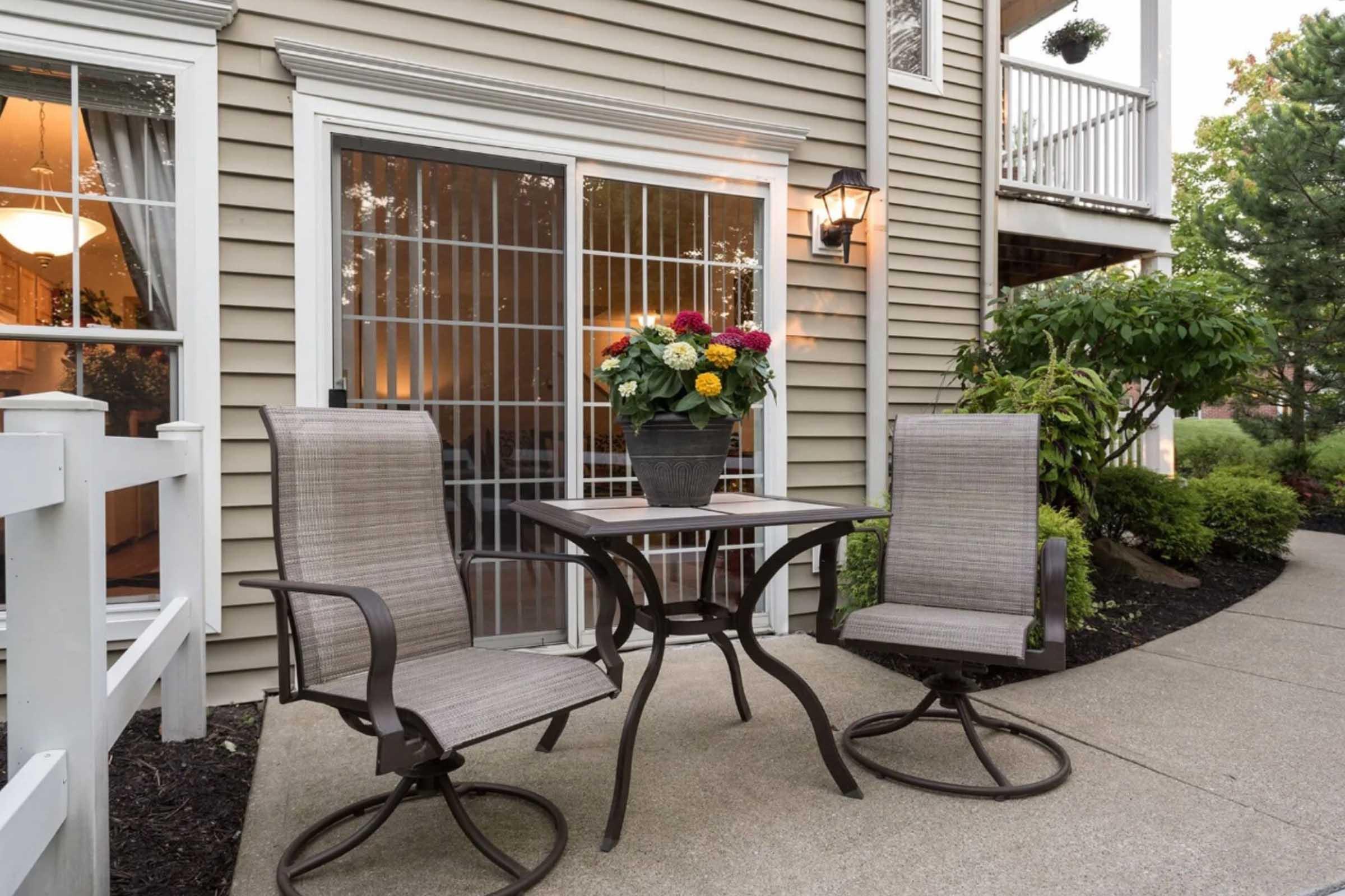 A cozy outdoor seating area featuring two swivel chairs and a small round table set up on a concrete patio. There's a potted flower arrangement on the table, and the patio is bordered by a white railing. The backdrop includes a house with sliding glass doors and windows, illuminated in the evening light.