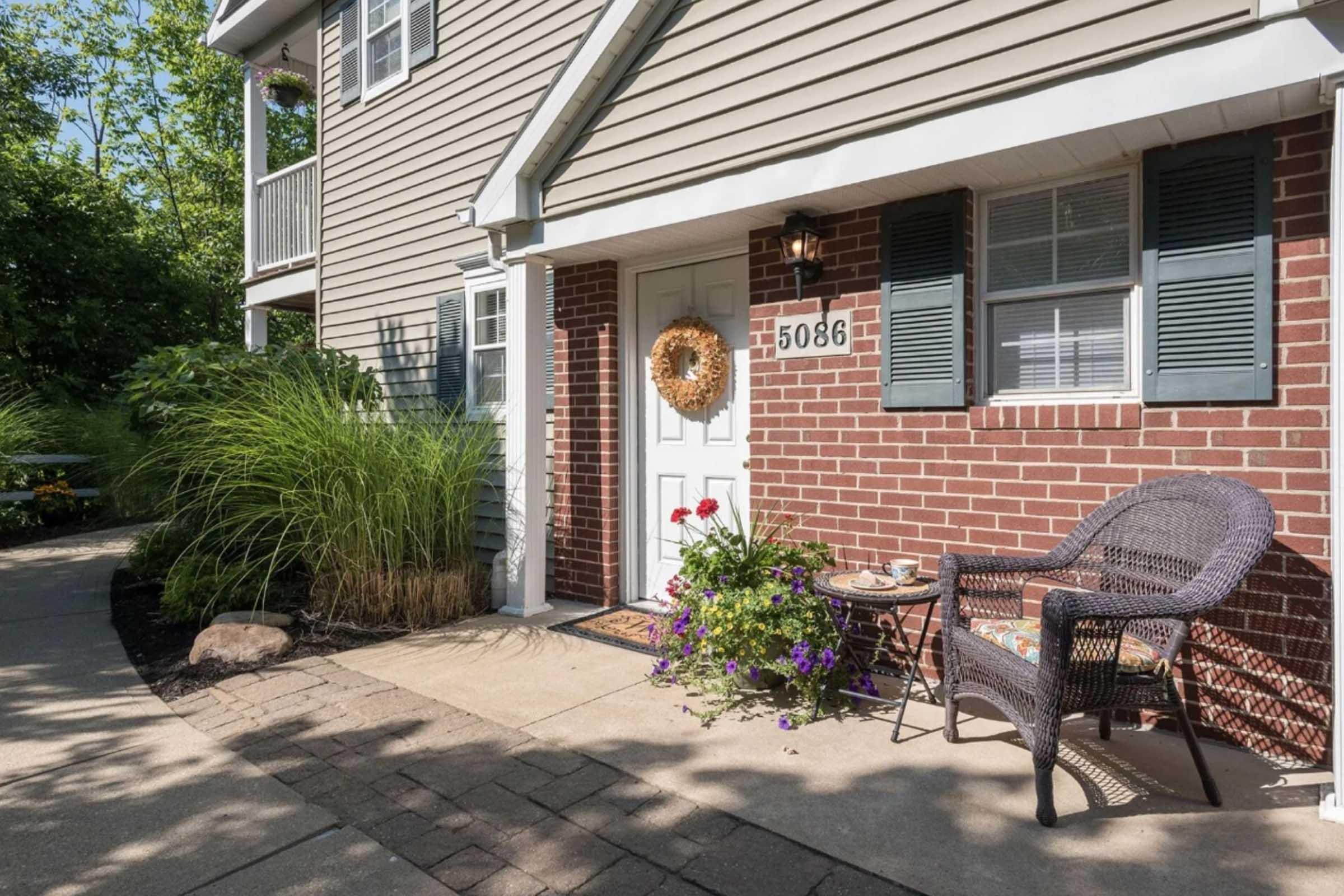 A welcoming entrance to a home featuring a brick façade, white door with a decorative wreath, and a wicker chair beside a table. Colorful flowers in pots and lush greenery enhance the inviting atmosphere, with a paved walkway leading up to the entryway.