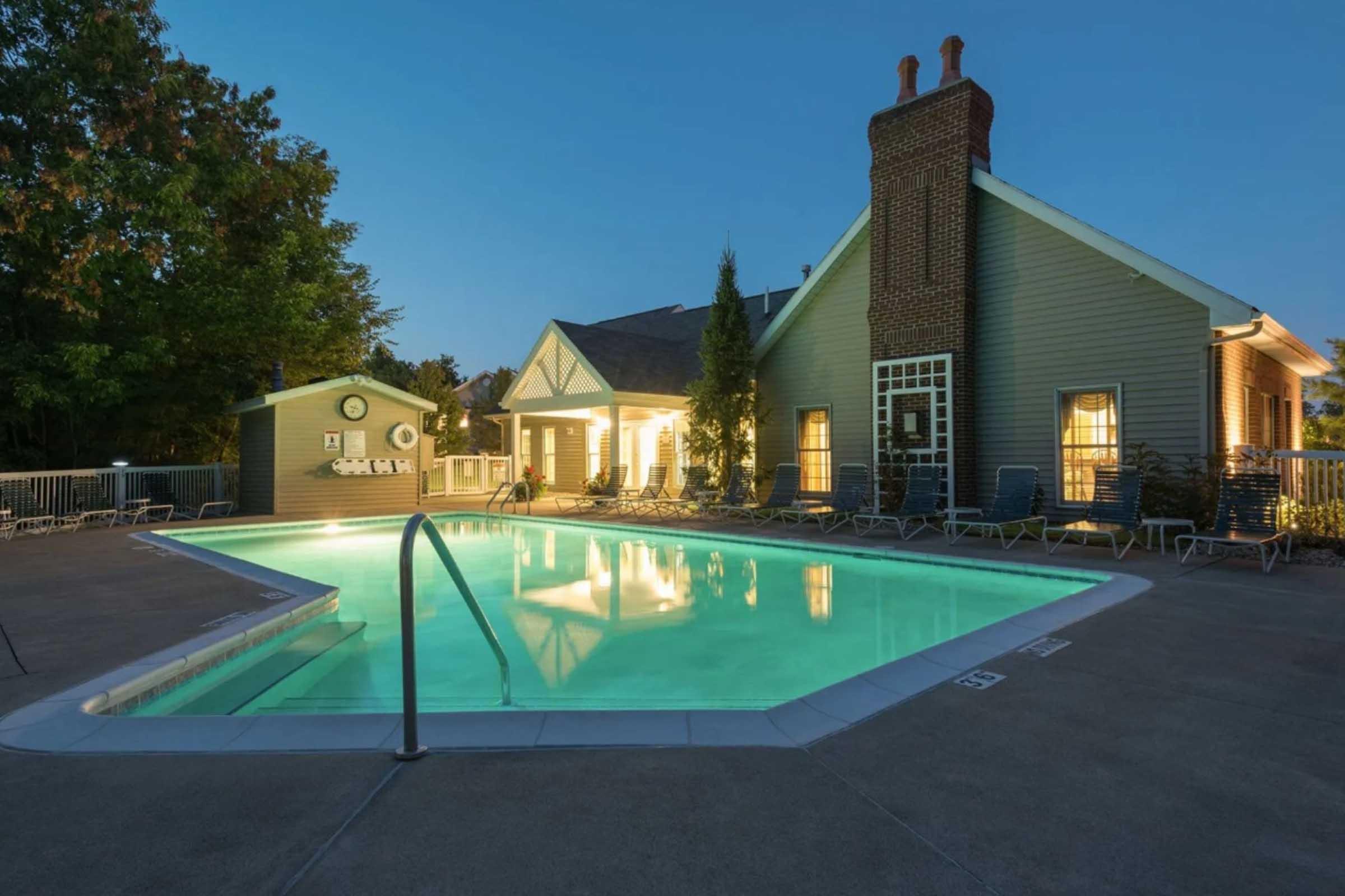 A tranquil pool area at dusk, featuring a well-lit, inviting swimming pool surrounded by lounge chairs. The buildings nearby have large windows, showcasing warm indoor lighting, and lush greenery adds to the serene atmosphere.