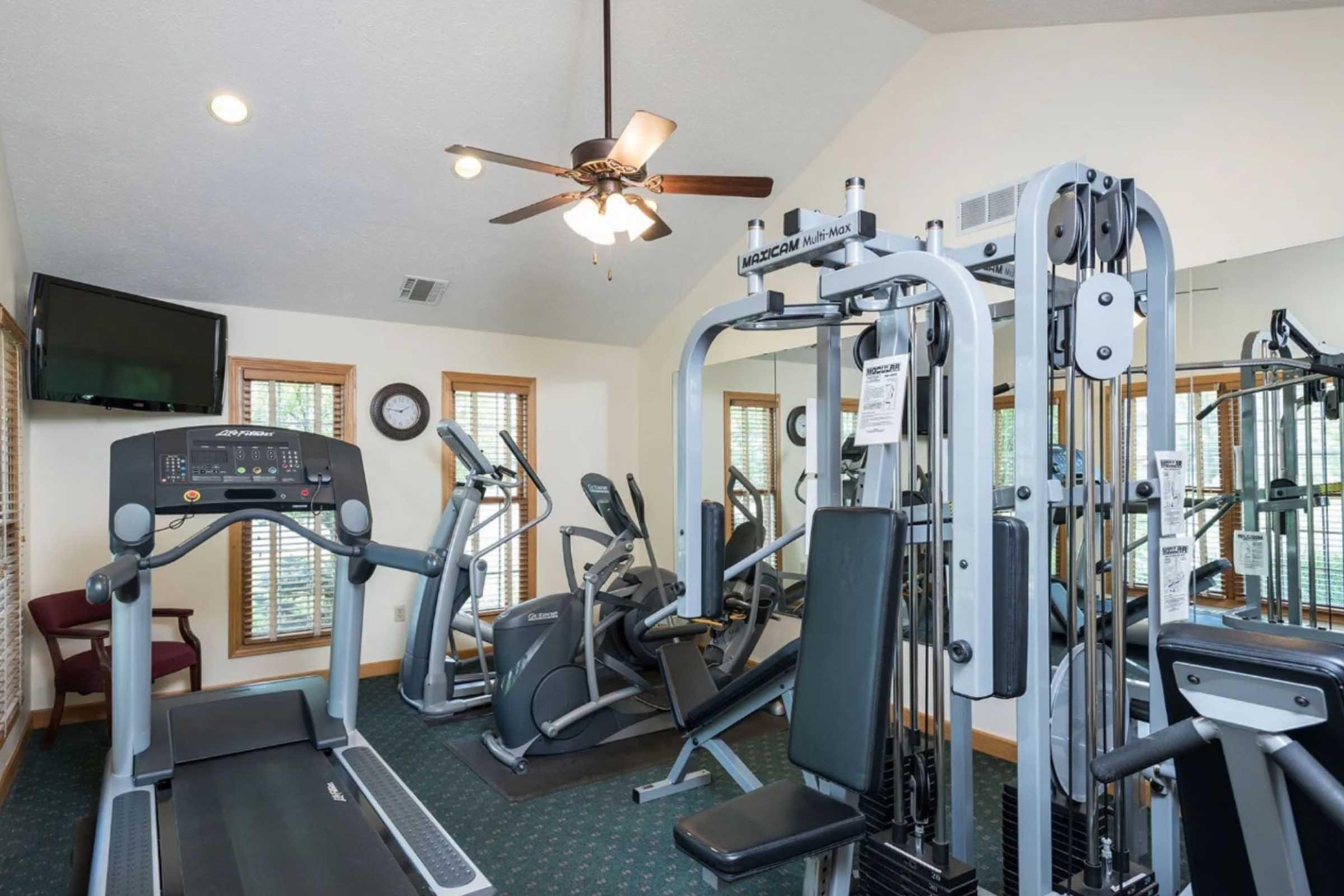 A well-equipped gym with various fitness machines, including a treadmill and strength training equipment. The room features large windows with natural light, ceiling fans, and wall clocks, creating a comfortable workout environment.