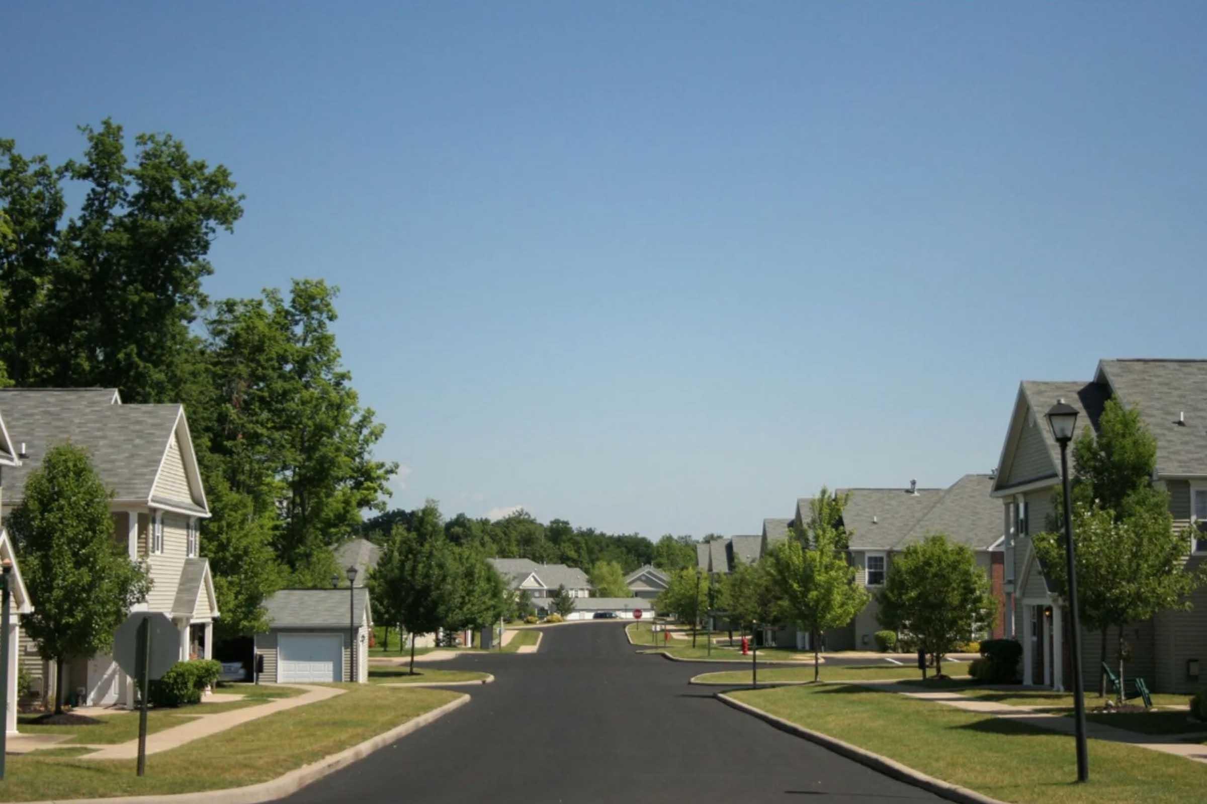 A quiet suburban neighborhood with well-maintained houses lining a paved street. Lush green trees and neatly manicured lawns are visible, under a clear blue sky. Streetlights and sidewalks enhance the peaceful residential atmosphere.