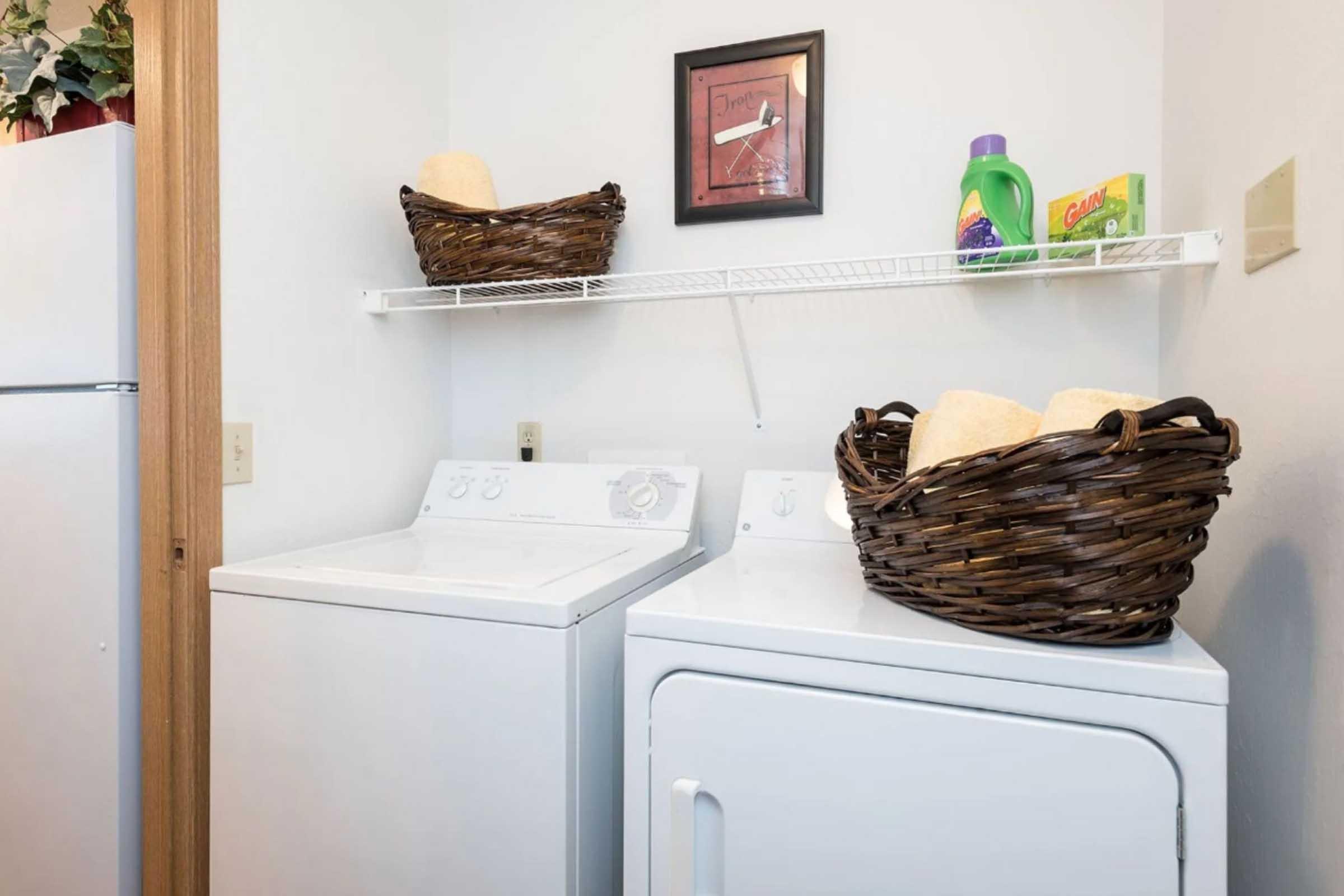 A laundry room featuring a white washing machine and dryer side by side. Above them, a wire shelf holds two wicker baskets filled with items, and a framed picture is on the wall. A bottle of laundry detergent is also visible on the shelf.