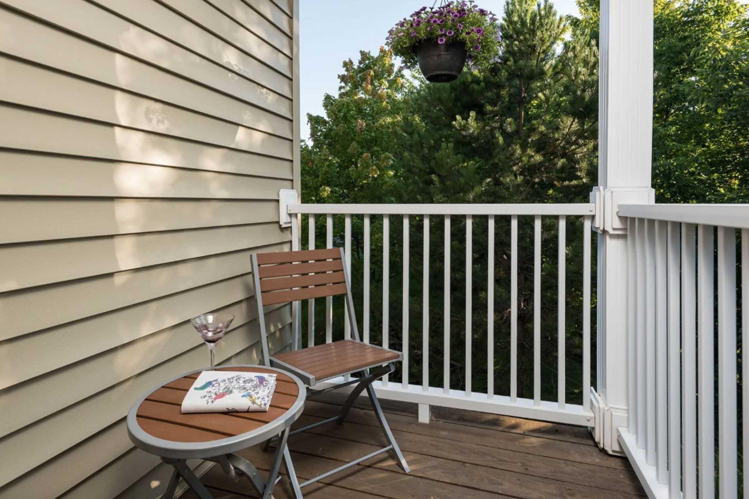 A small balcony featuring a wooden folding chair and a round table with a glass of drink. A decorative plant hangs above, and greenery is visible in the background, creating a serene outdoor space.