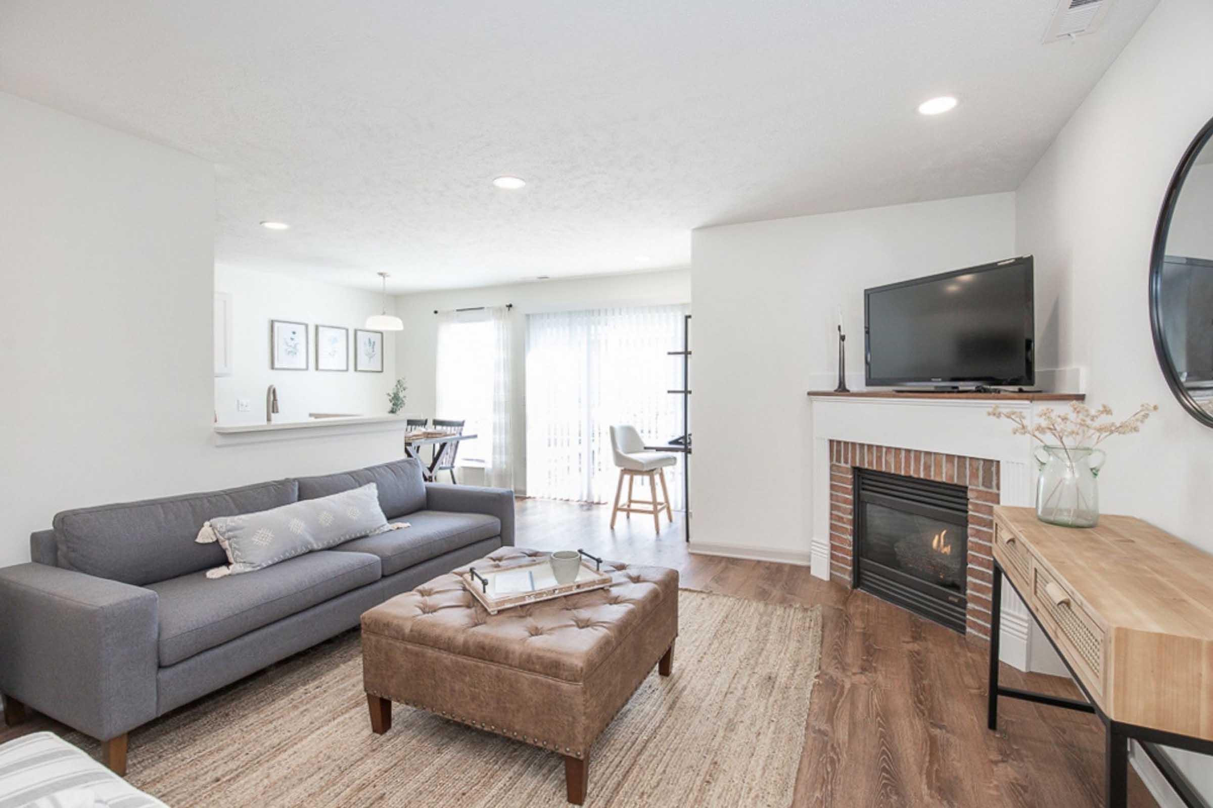A modern living room featuring a gray sofa with decorative pillows, a brown ottoman, a wooden side table, and a fireplace. There's a television mounted on the wall, and large windows with sheer curtains allow natural light to fill the space. A small dining area is visible in the background.