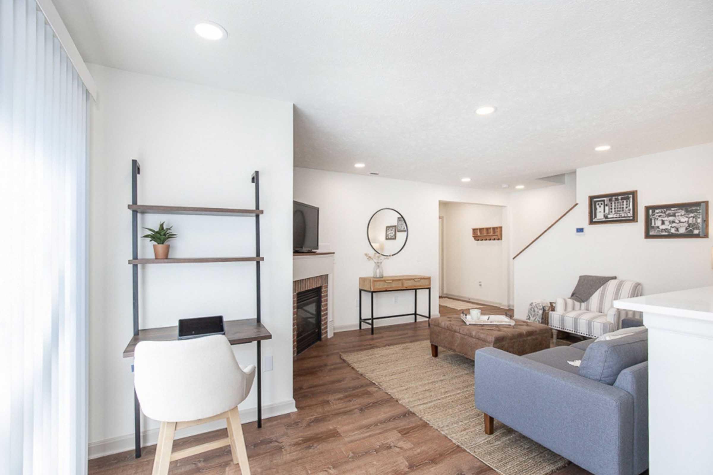 Modern living room with a cozy seating area featuring a gray sofa, striped chairs, and a wooden coffee table. A small desk with a chair and a plant is near a fireplace. Wall-mounted TV and round mirror enhance the decor. Light-colored walls and wooden flooring create a bright, inviting atmosphere.