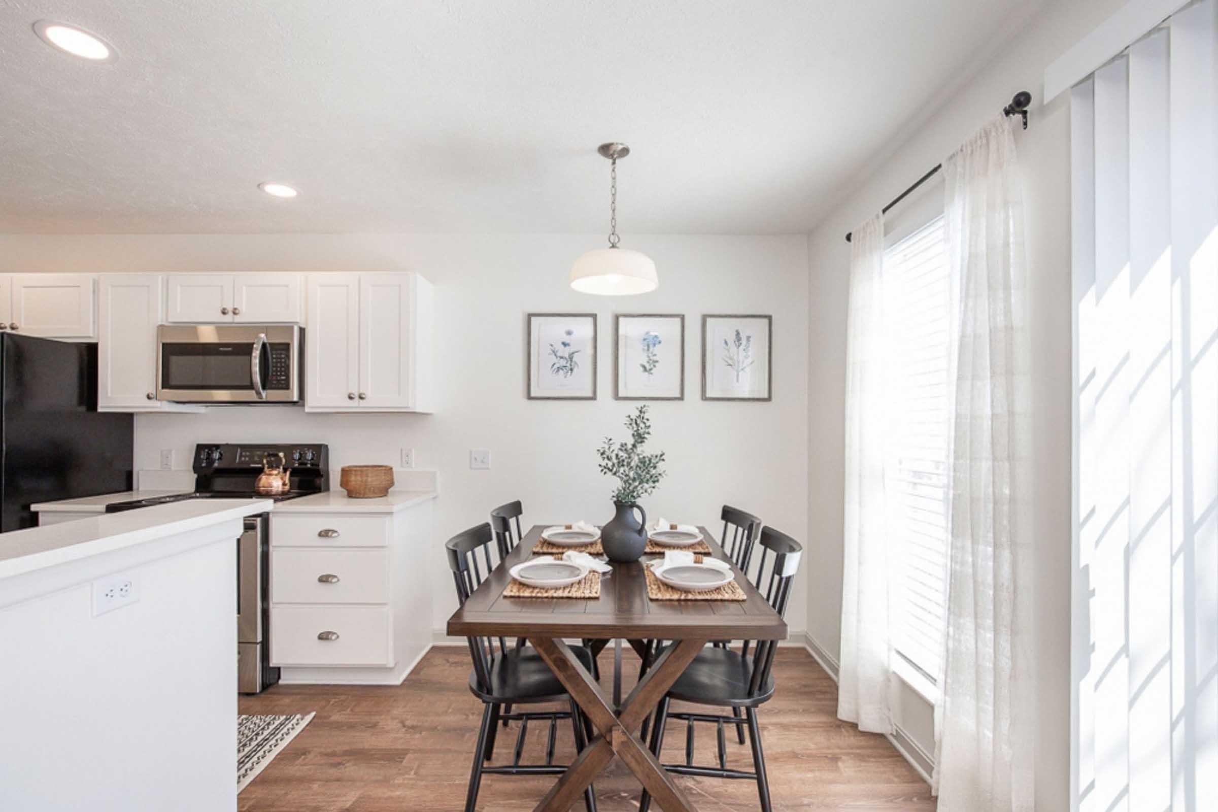 A modern kitchen with white cabinetry and stainless steel appliances, featuring a wooden dining table set for four. Decorative plates and a vase with greenery are on the table. Light streams in through large windows adorned with sheer curtains, complemented by framed botanical art on the wall.