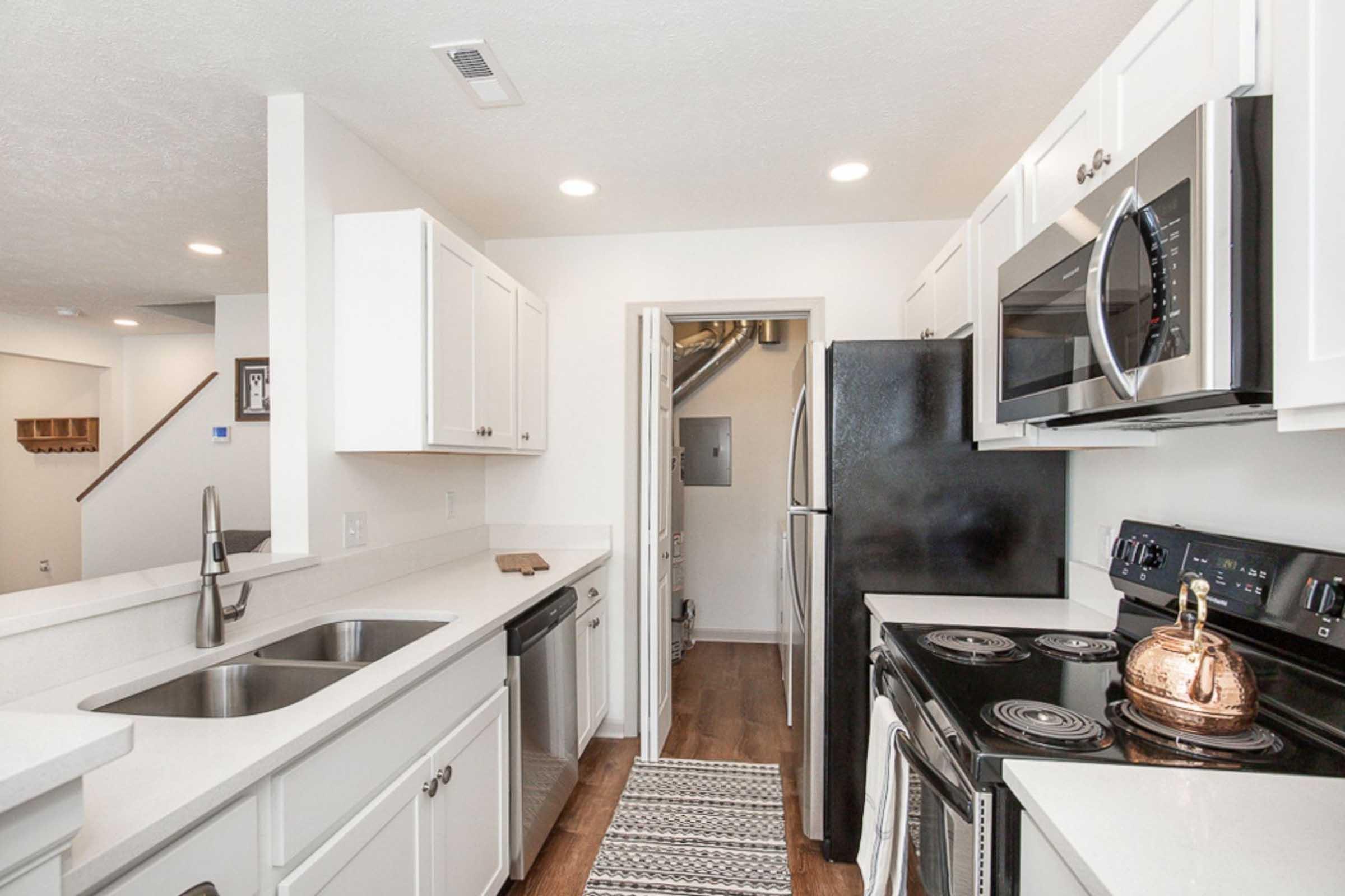 Modern kitchen with white cabinetry, stainless steel appliances including a microwave and stove, a black refrigerator, and a double sink. The countertops are light-colored and there's a decorative rug on the floor. A doorway leads to another room, and overhead lighting brightens the space.