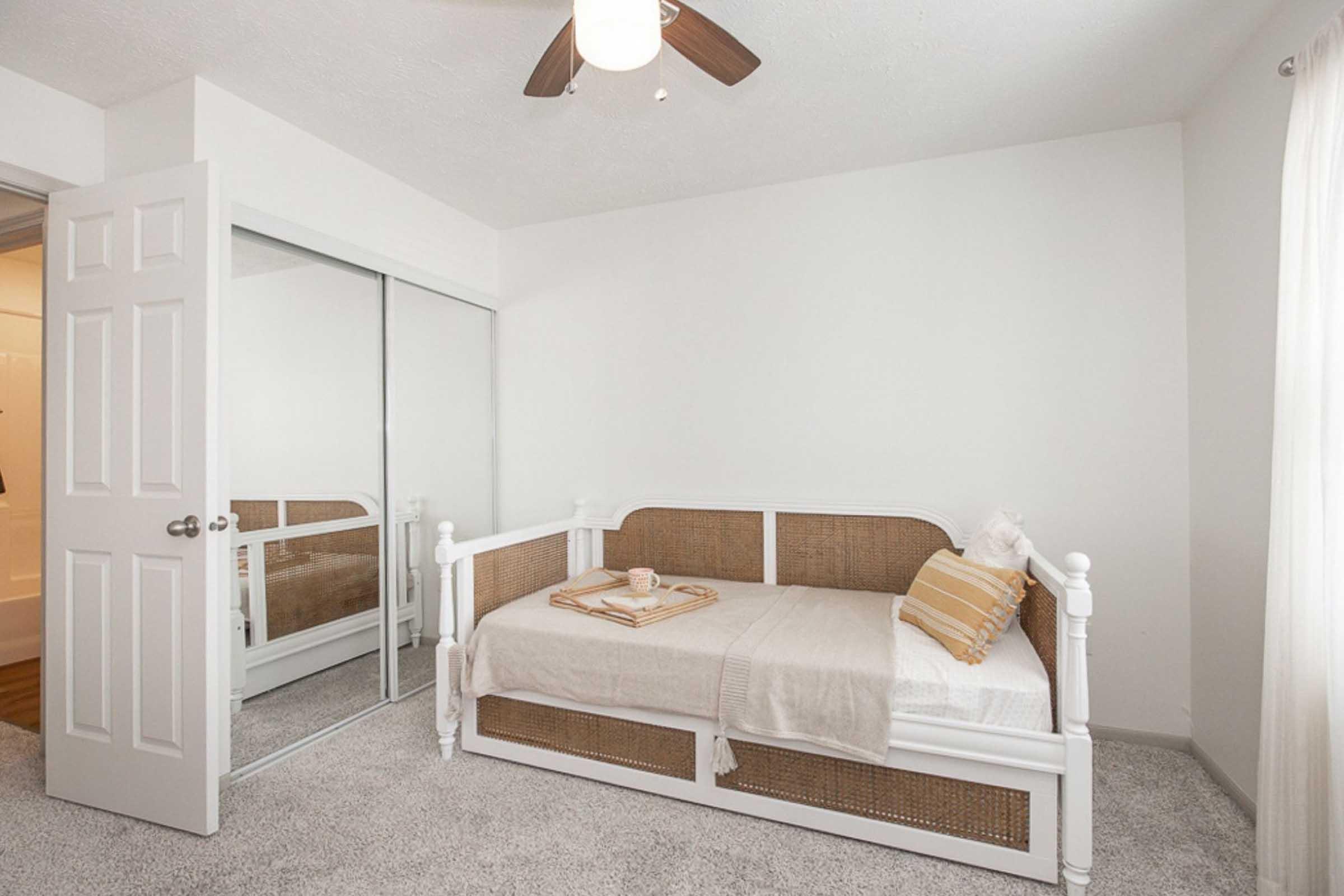 A cozy bedroom featuring a white daybed with a beige and white striped pillow, a light-colored throw blanket, and a tray with a cup on it. There's a mirror closet door on one side and a ceiling fan above, with light-colored walls and carpet, creating a calm and inviting atmosphere. Bright natural light enters through a window with sheer curtains.