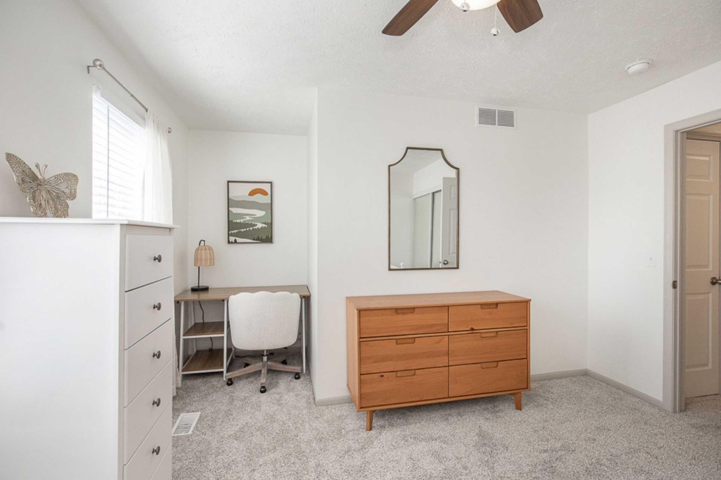 A minimalist bedroom featuring a light color scheme. There is a wooden dresser with multiple drawers, a desk with a chair, and a framed artwork on the wall. A mirror hangs above the dresser, and a small lamp sits on the desk. The room has carpet flooring and a ceiling fan.