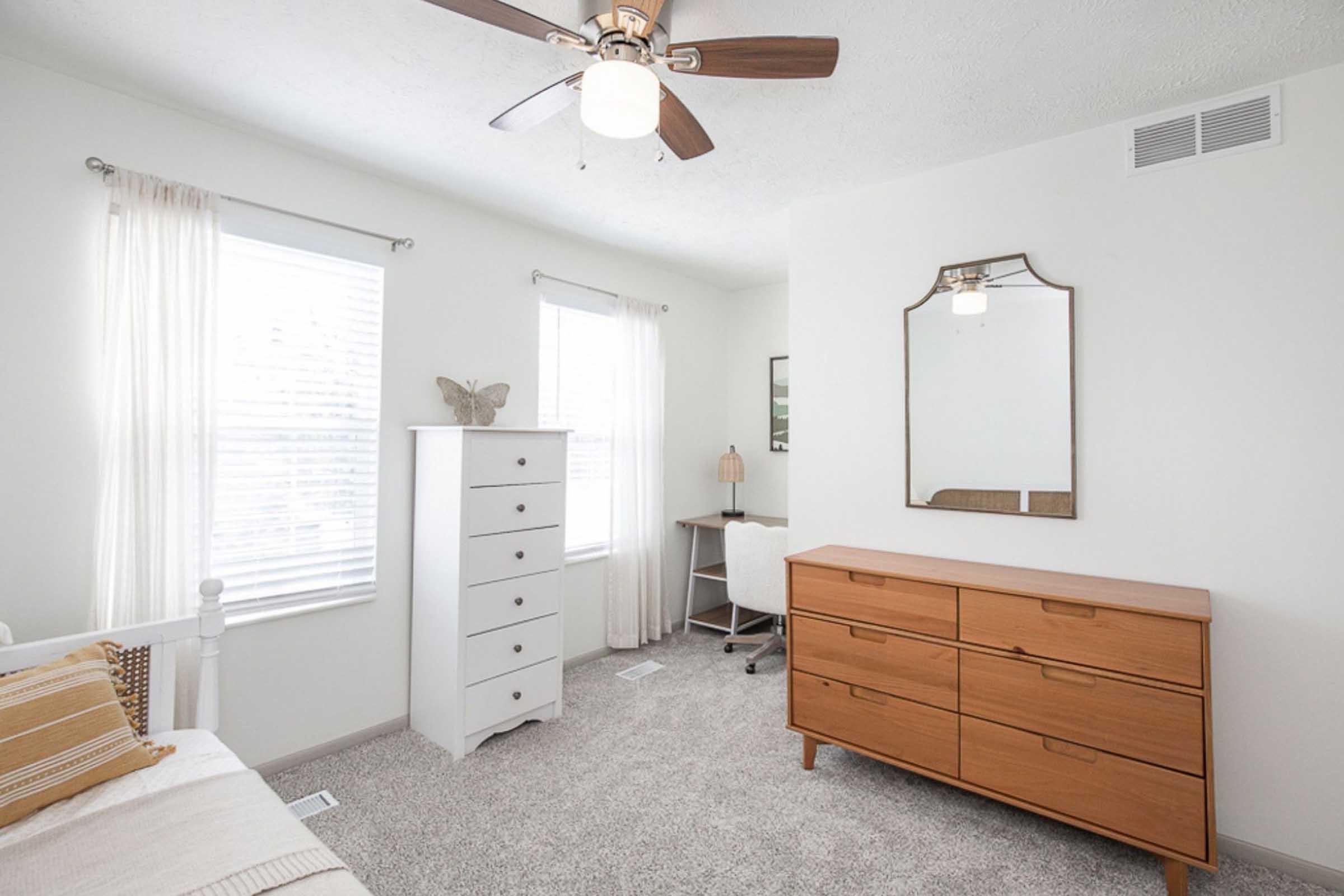 A bright and airy bedroom featuring a white bed with decorative pillows, a white dresser, a wooden dresser with multiple drawers, a small desk with a lamp, and two windows with beige curtains. A ceiling fan adds a modern touch, and a mirror hangs on the wall opposite the bed. The carpeted floor enhances the cozy atmosphere.