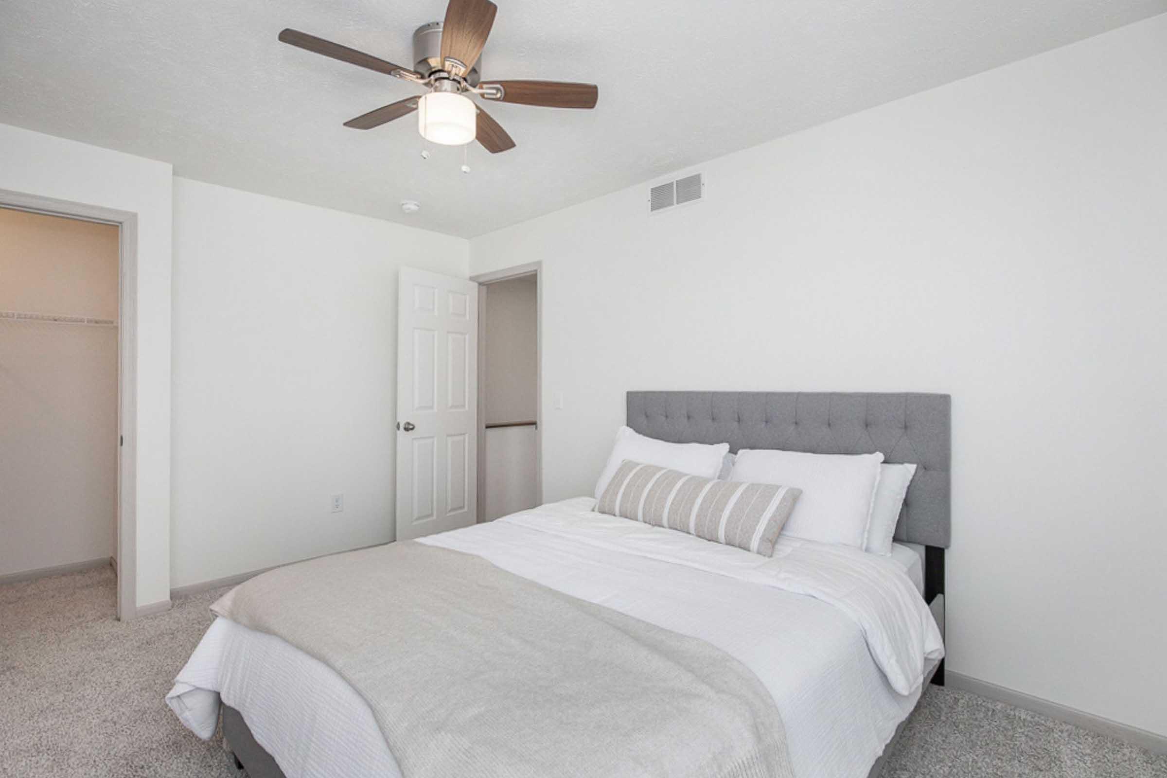 A cozy bedroom featuring a grey upholstered bed with white bedding and decorative pillows. There is a light-colored ceiling with a modern ceiling fan and a nearby closet door. The walls are painted white, and the floor is covered with plush carpet, creating a comfortable and inviting atmosphere.