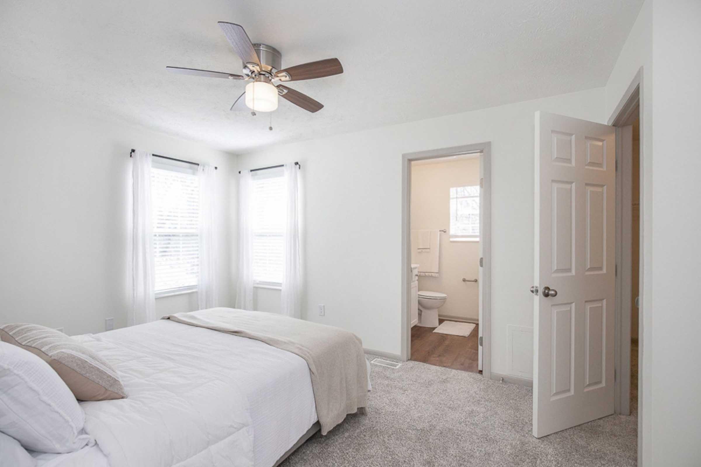 Bright and airy bedroom featuring a queen-sized bed with decorative pillows, a ceiling fan, and large windows with sheer curtains. A doorway leads to a bathroom with light-colored walls and a visible toilet. The room has a cozy carpeted floor, creating a comfortable and inviting atmosphere.