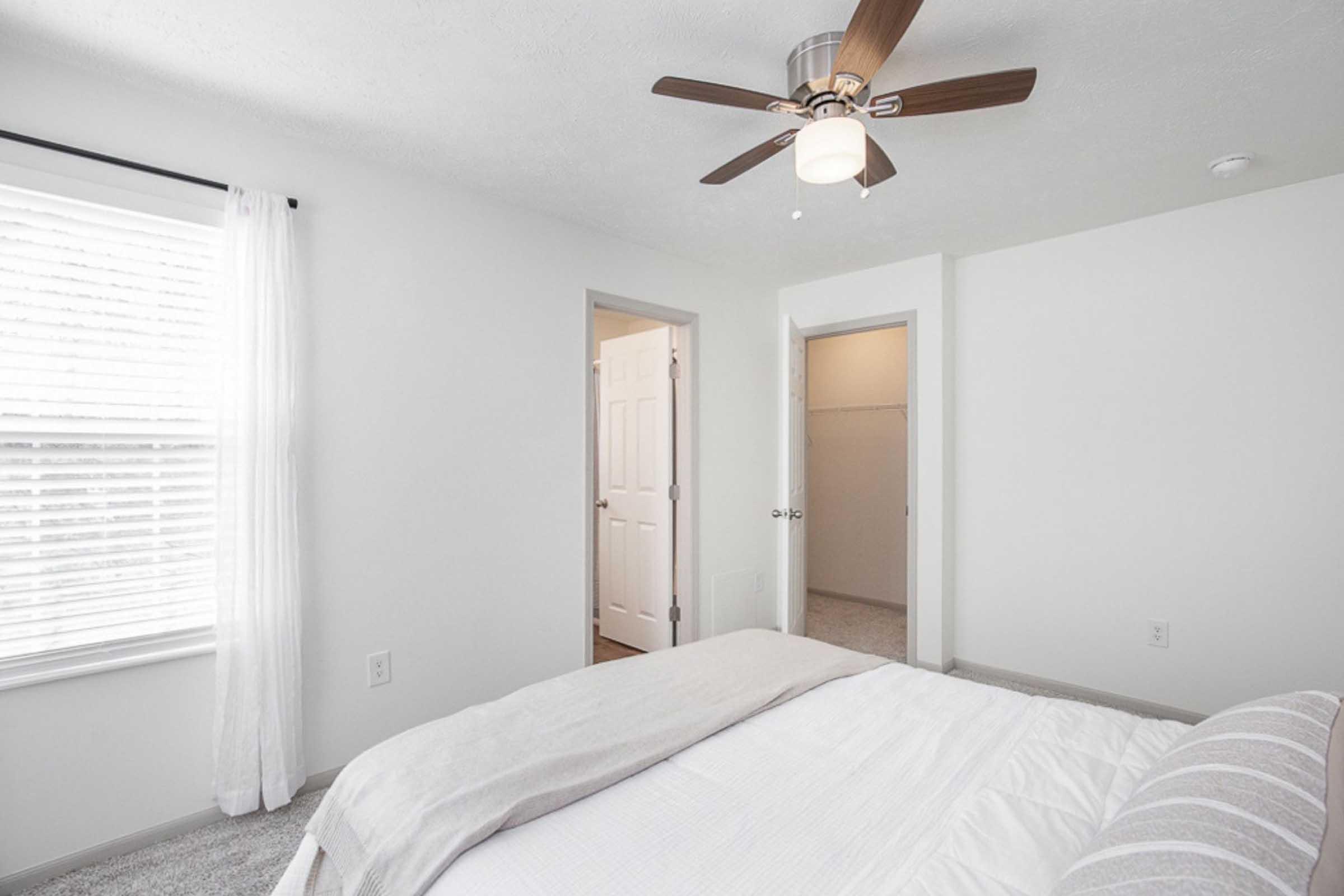A well-lit bedroom featuring a queen-sized bed with a white comforter, a ceiling fan with wooden blades, and light-colored walls. Natural light filters through a window with sheer white curtains. Doors lead to a closet and another room. The carpeted floor adds warmth to the space.