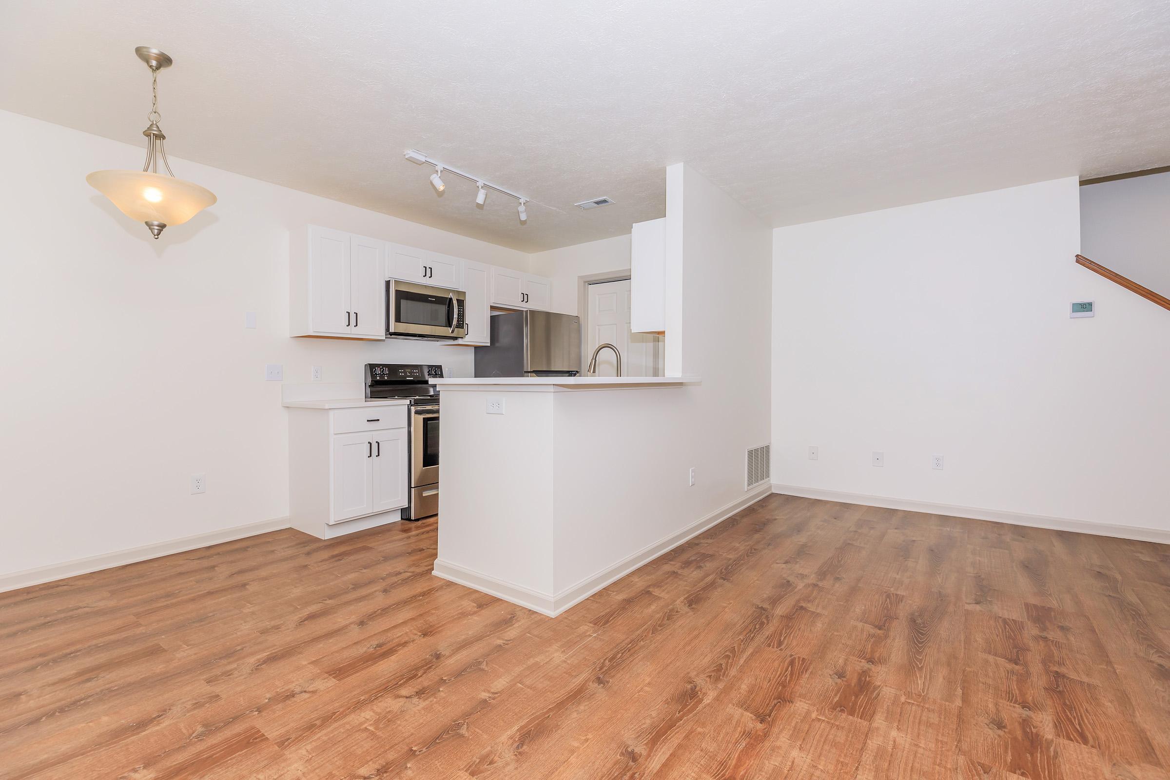 A modern kitchen and living area featuring white cabinetry, stainless steel appliances, and hardwood-style flooring. The space is well-lit with natural light and includes a pendant light fixture above the kitchen island. There is an open layout with a staircase visible in the background.
