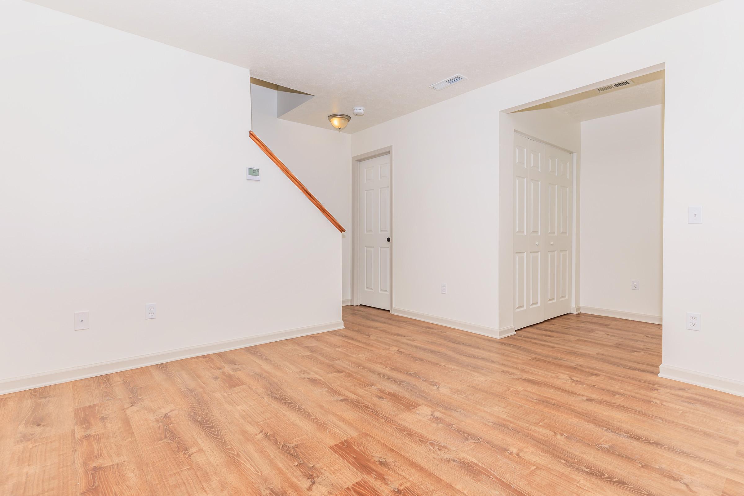 A spacious, empty room with light-colored walls and hardwood flooring. A staircase is visible in the background, along with a doorway leading to another room. The overall ambiance is bright and clean, making it suitable for various uses, such as a living room or office space.