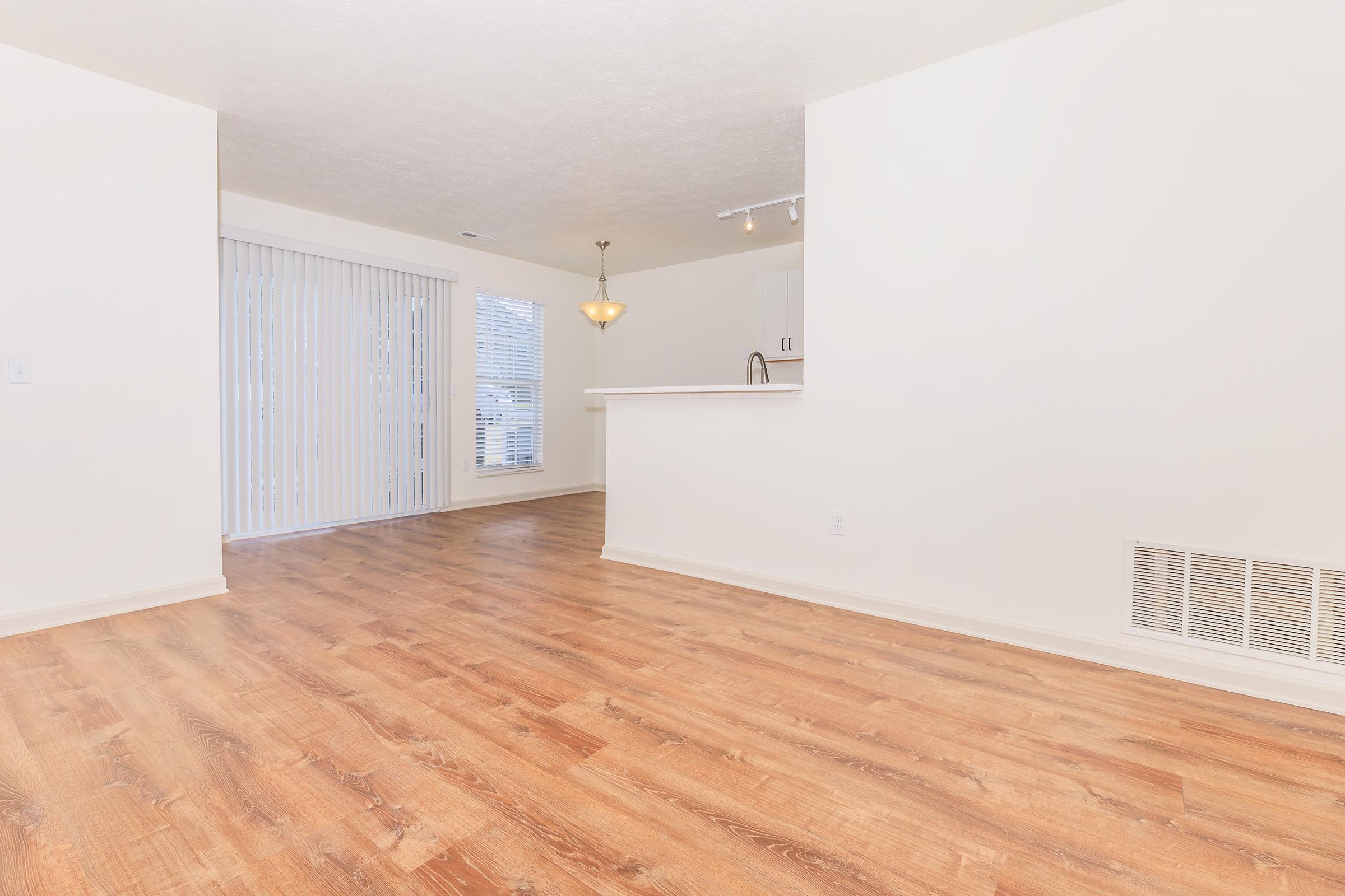 Spacious living area with light-colored walls and wooden flooring. A large window with blinds allows natural light, and there is a partitioned kitchen area with a bar counter in the background. The space appears clean and inviting, ready for furniture arrangement.