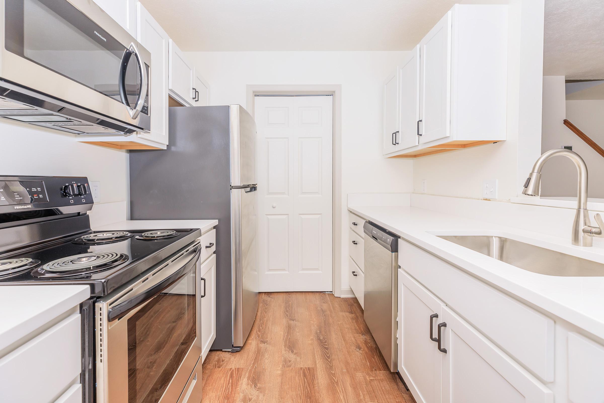 Modern kitchen featuring stainless steel appliances including a stove, refrigerator, and dishwasher. The cabinets are white, and the countertops are light-colored. A sink with a sleek faucet is visible, and the flooring is a warm-toned wood. The layout is clean and contemporary, with a doorway leading to another room.