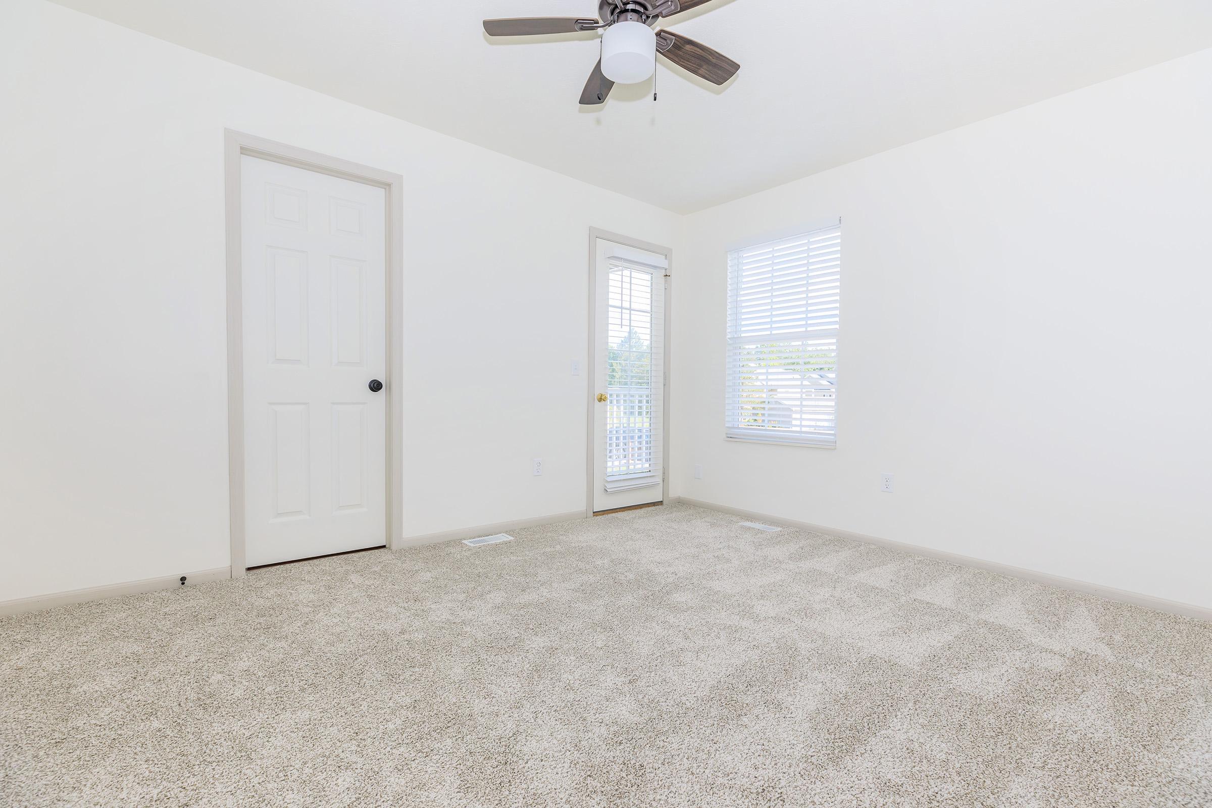 A well-lit, empty room featuring light-colored walls, a ceiling fan, and soft beige carpet. There is a closed white door and a set of glass doors with blinds leading to an outdoor balcony, along with a window allowing natural light. The space appears clean and ready for furnishing.