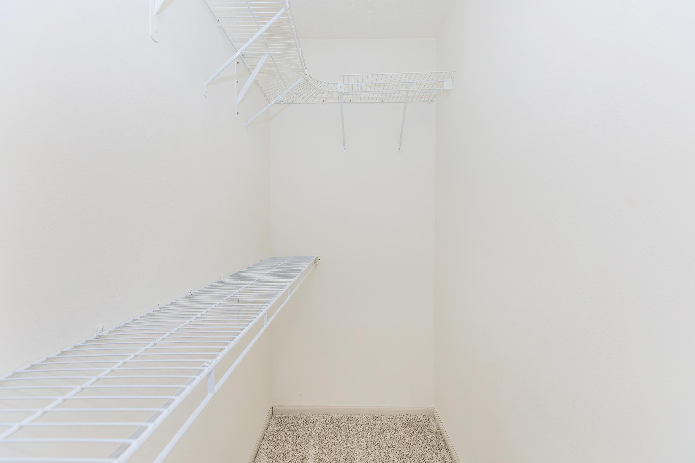 Empty closet featuring white wire shelving mounted on light-colored walls. The floor is carpeted in a neutral tone. The space is bright and uncluttered, emphasizing the minimalist design of the storage area.